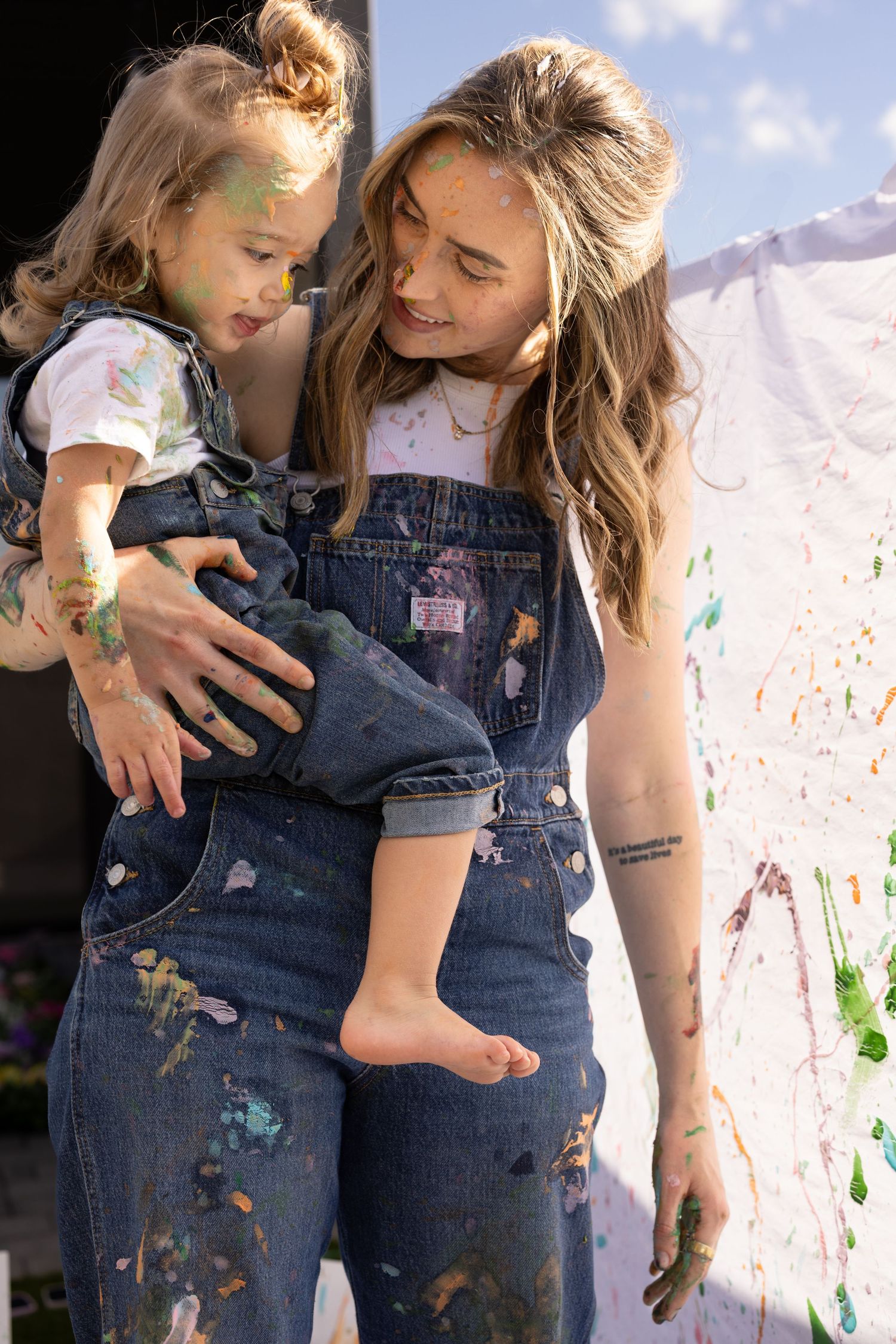 Mother holding daughter painting photoshoot in Arizona, in denim overalls, covered in paint, smiling. mommy and me shoot