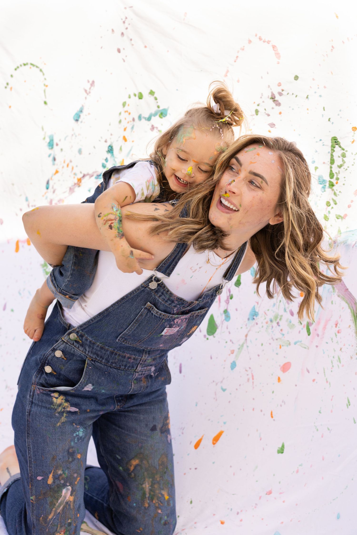 tender mother daughter moment in painting family mommy and me photoshoot. laughing and covered in paint, in denim overalls and white t-shirt