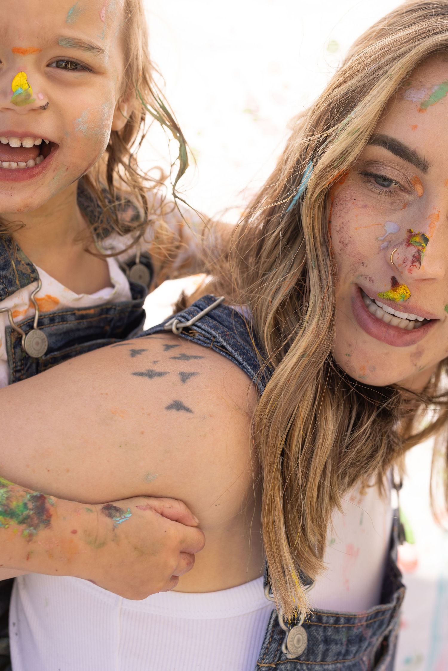 mother giving daughter piggyback ride during a painting photo session in a mommy and me photoshoot. covered in splattered paint and laughing