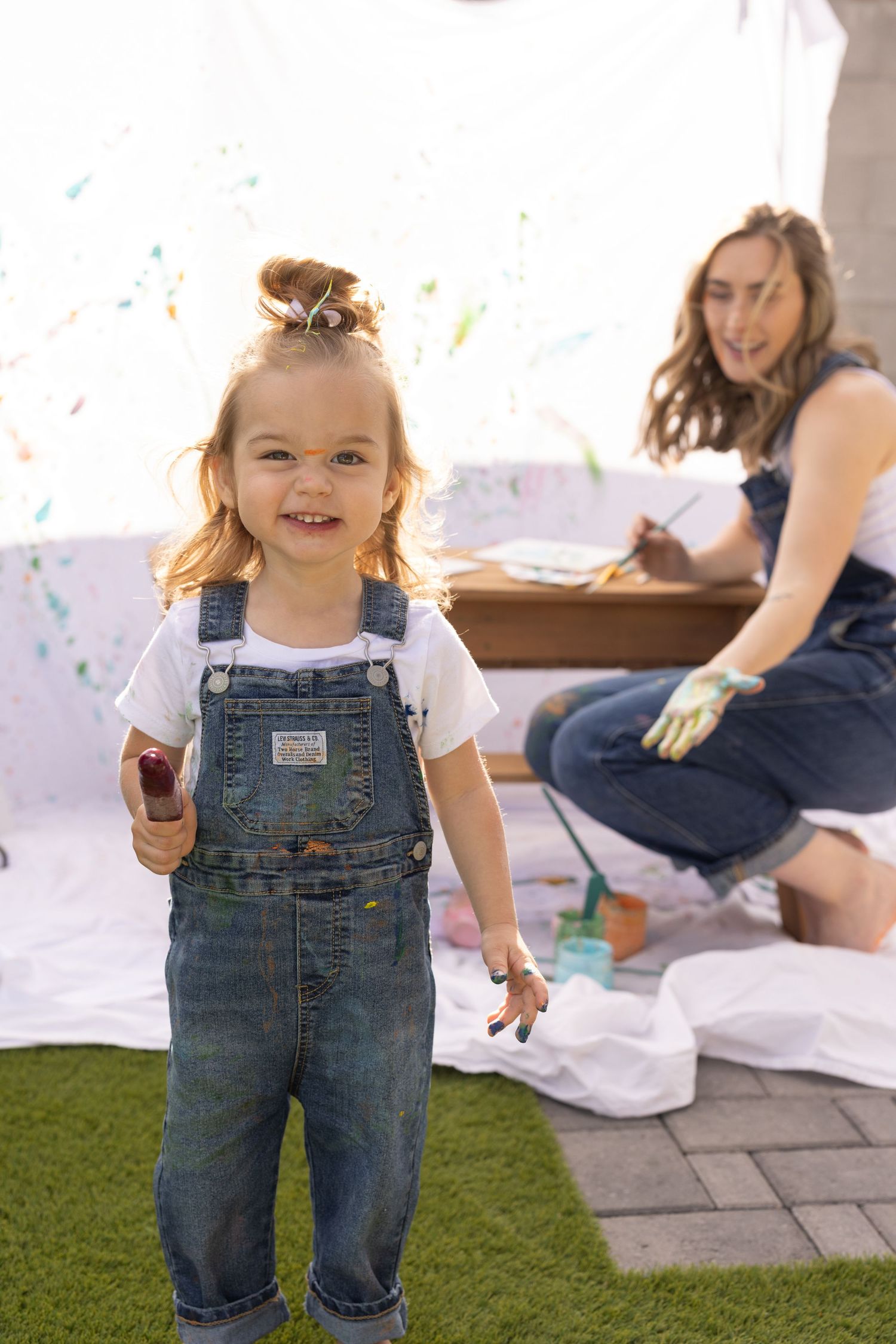 girl holding popsicle painting with mom in photoshoot, white sheet background. food colored yogurt used as safe non-toxic paint alternative