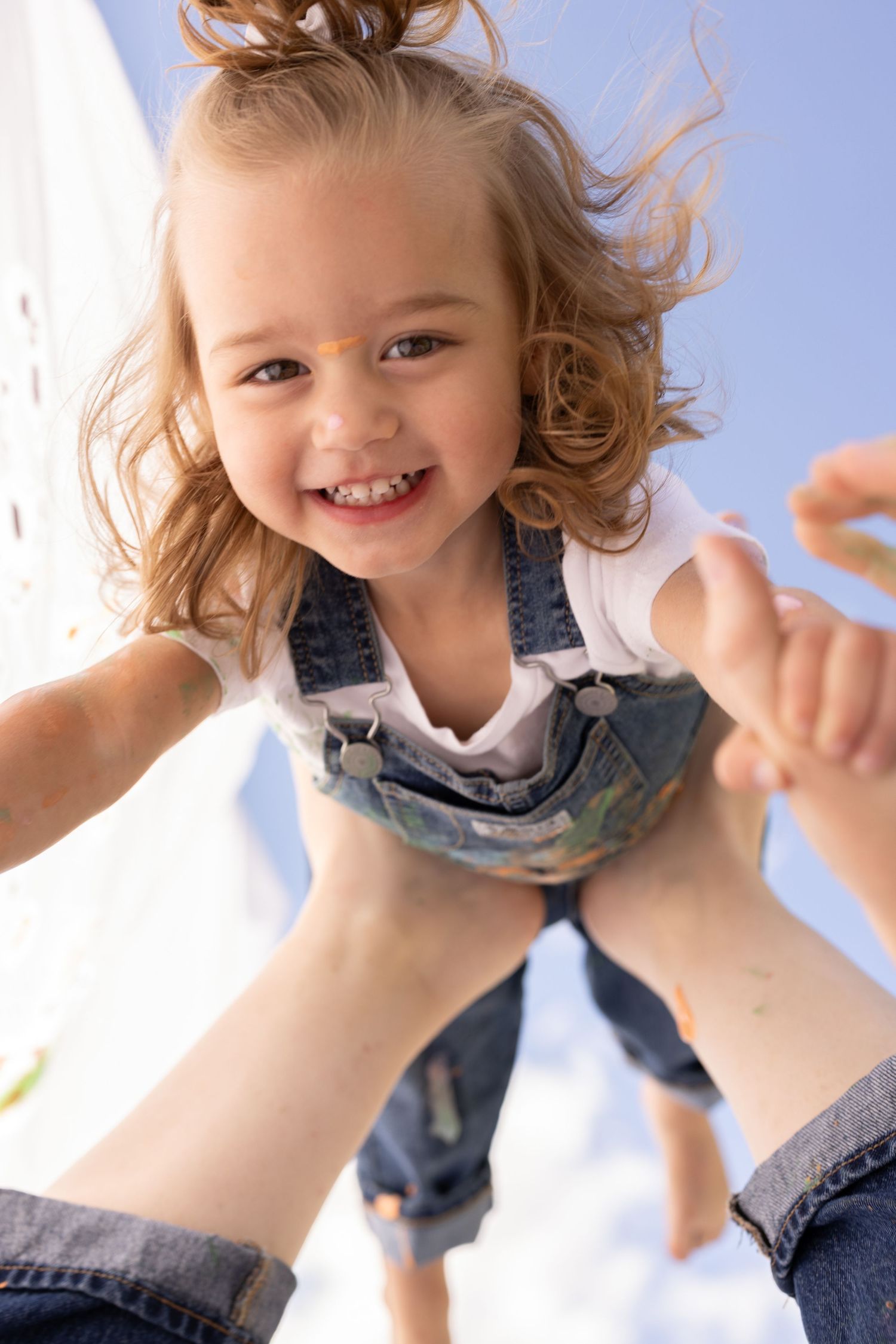 mother tossing daughter in the air blue skies in denim overalls and white t-shirt on a mommy and me painting photoshoot in Phoenix, AZ