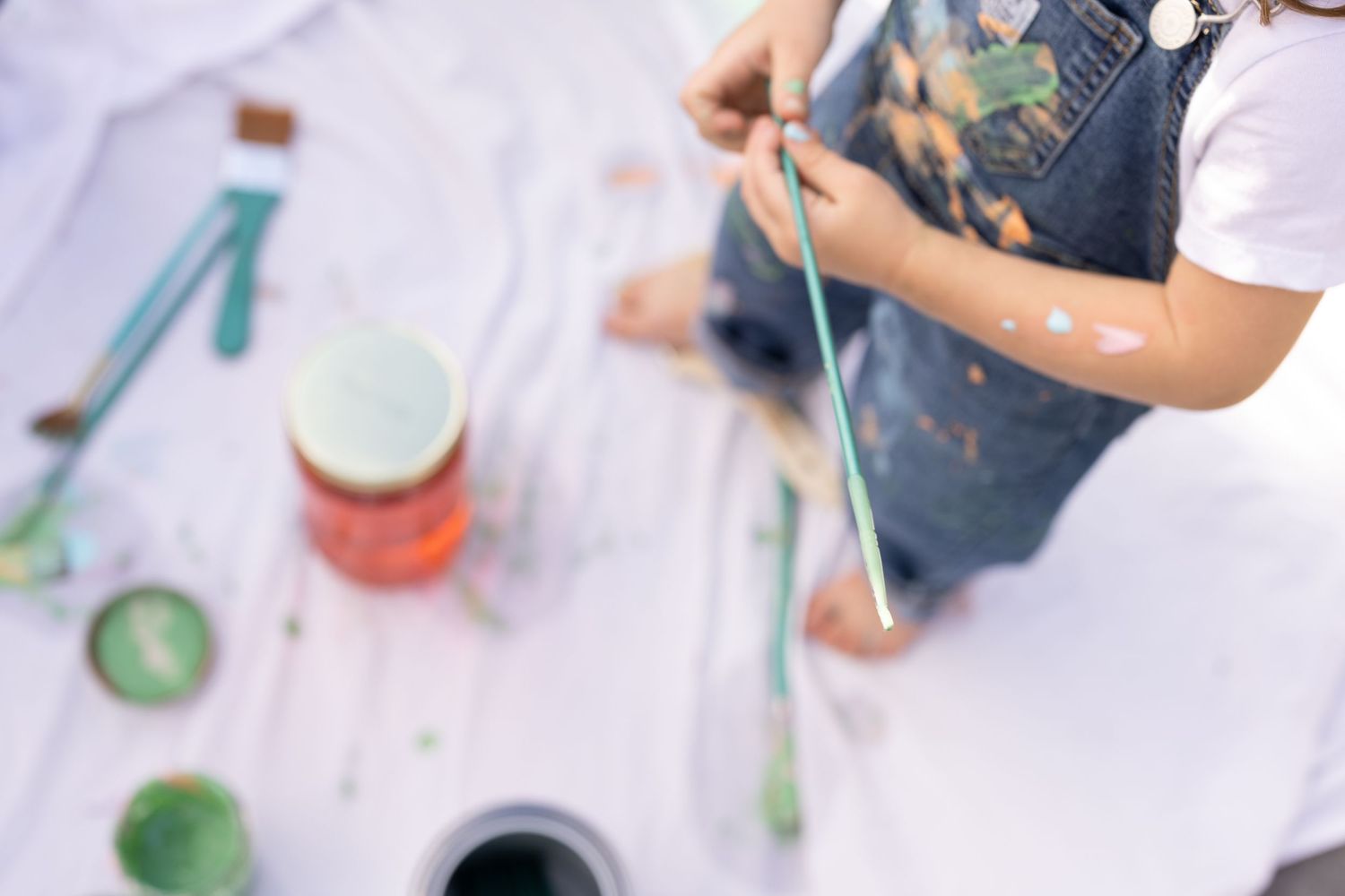 child holding paint brush, painting in mommy and me photo session. walking over white sheet and wearing overalls with white t-shirt