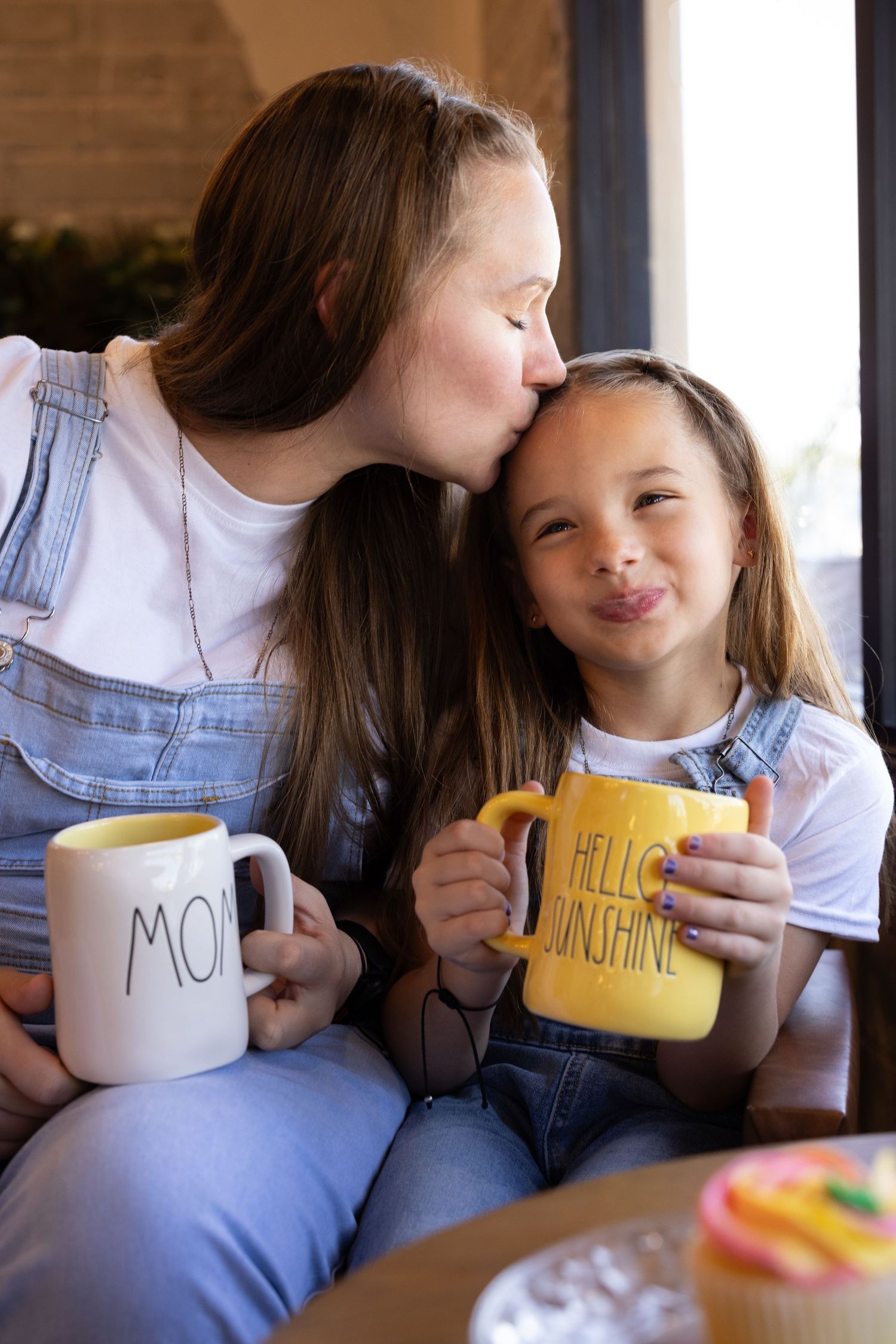 Mother kissing daughter on forehead, holding mugs wearing denim overalls on mommy and me family shoot at a local coffee shop in Phoenix, AZ