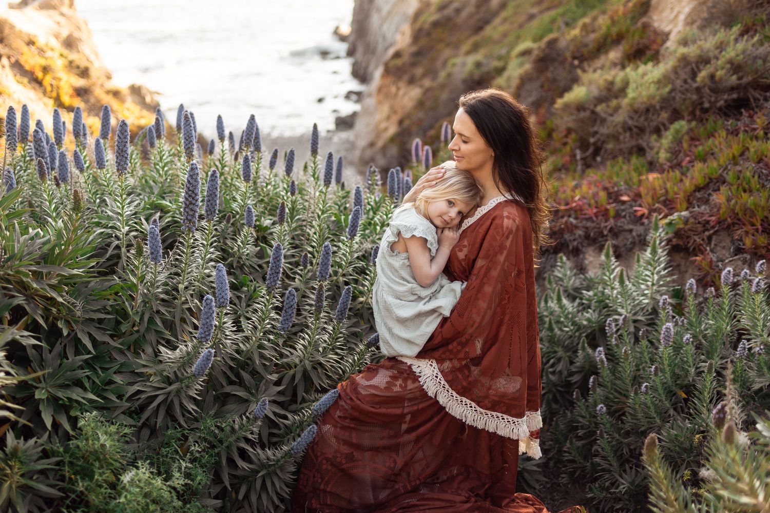 A mother in a red dress holds her child among purple wildflowers on a scenic coastal cliffside.