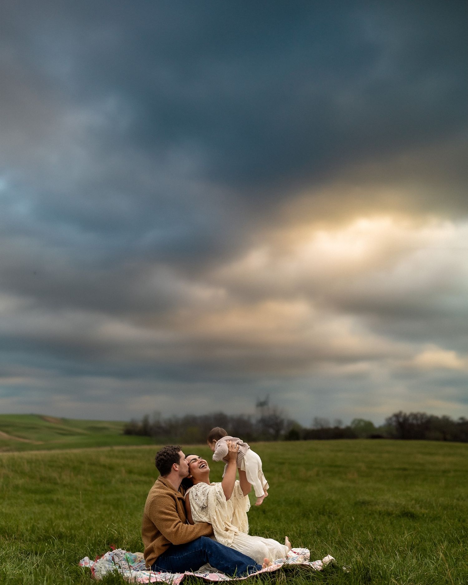 Family sitting on blanket in green field holding baby up under dramatic cloudy sunset sky.