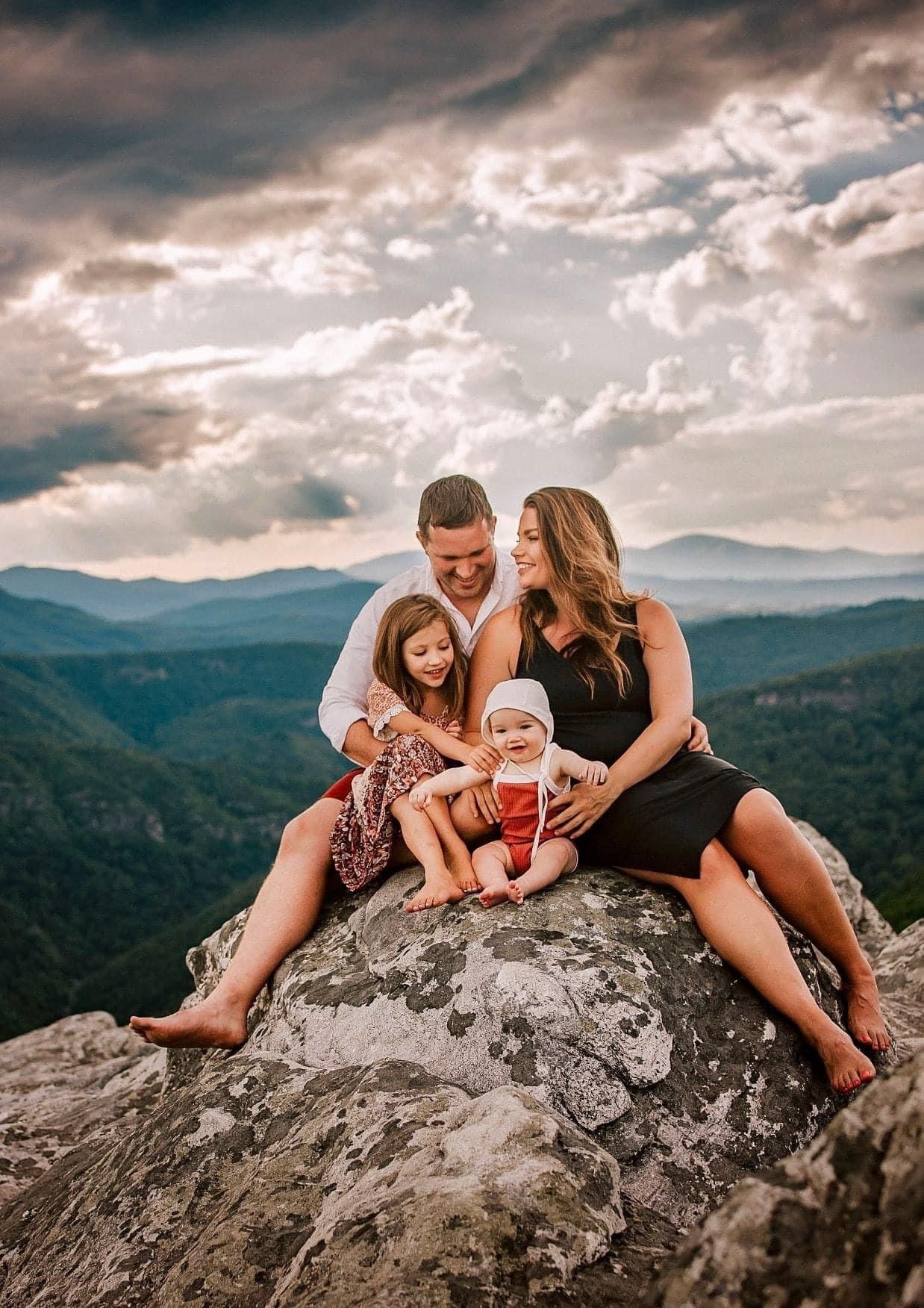 Family of four sitting on a rocky mountain overlook with scenic valley and dramatic cloudy sky backdrop.
