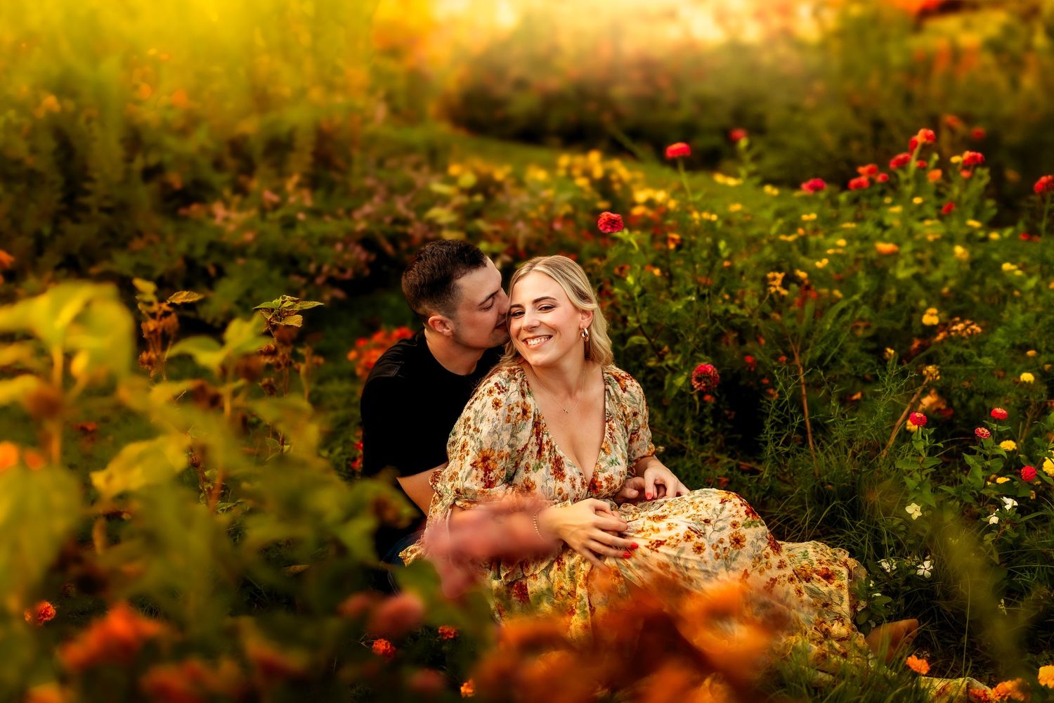 Couple sitting in a vibrant flower field, woman in floral dress smiling as man kisses her cheek at golden hour.