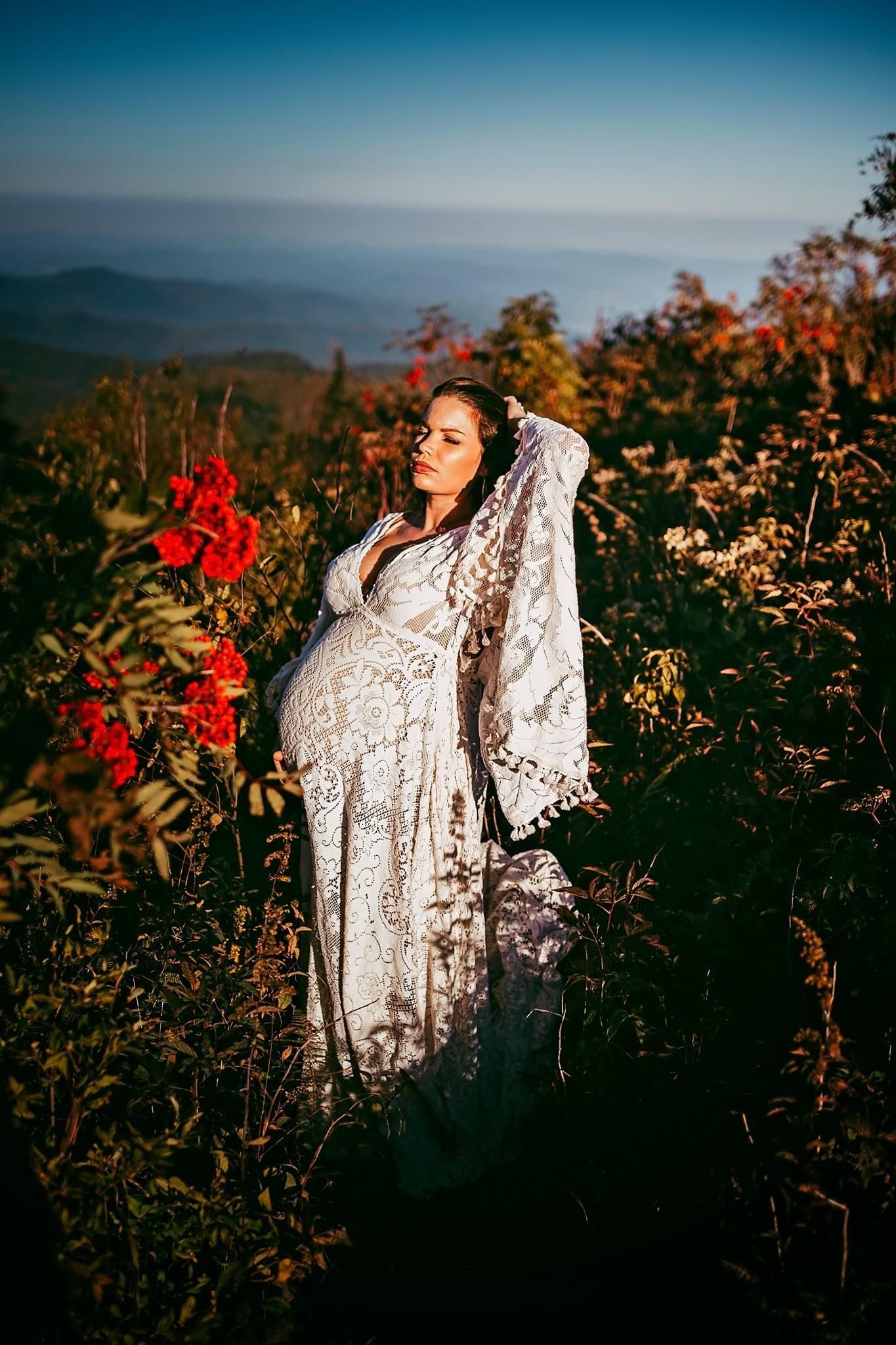 Pregnant woman in white lace dress standing among wildflowers on a mountain overlook at golden hour.