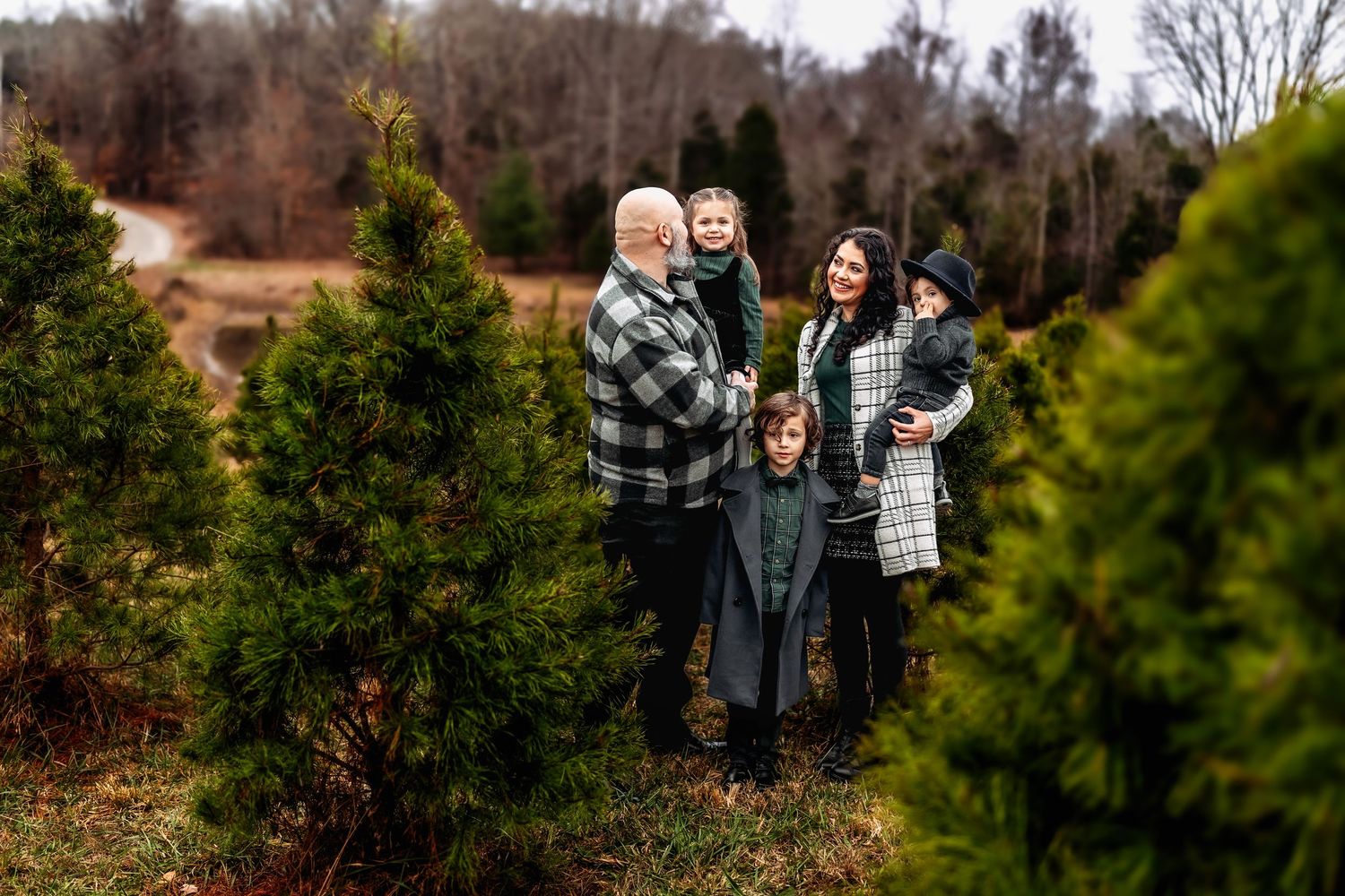 Family of five smiling together among evergreen trees at a Christmas tree farm in winter.
