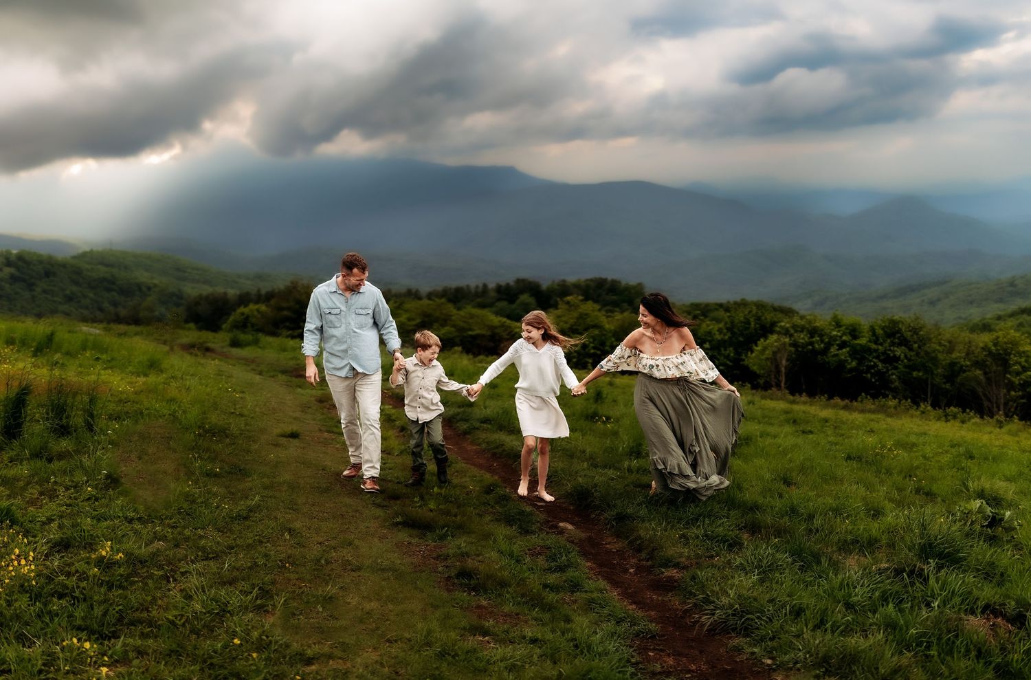 Family of four holding hands walking on a green mountain trail with dramatic stormy skies and misty peaks behind them.