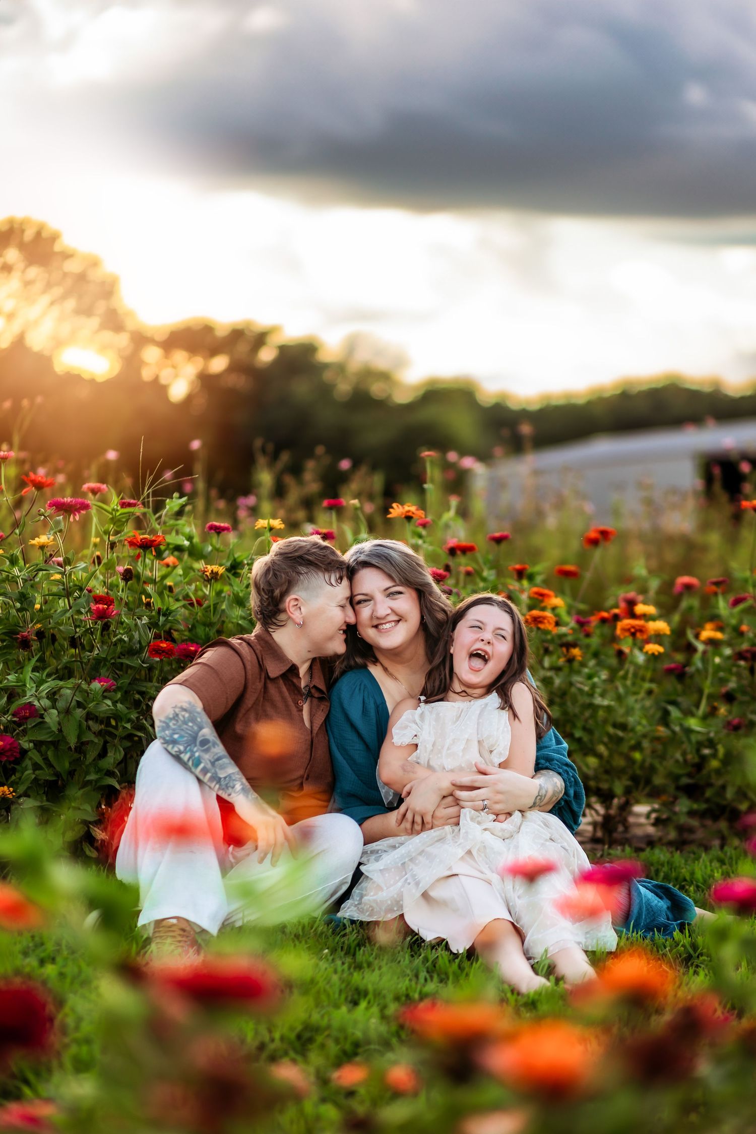 Family sitting in colorful flower field at golden hour, parents kissing laughing child among red and orange zinnias.