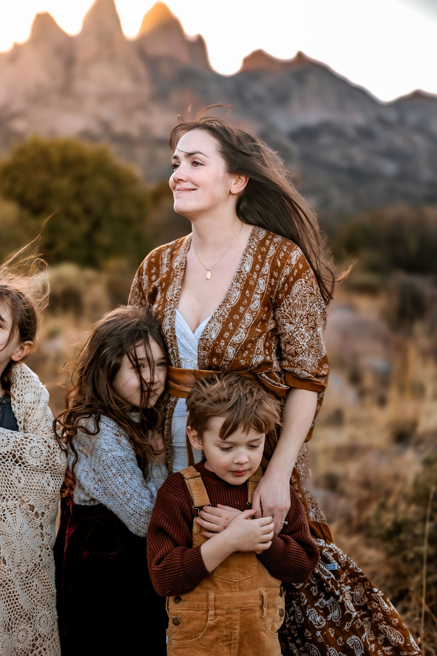 A smiling woman in a boho dress stands with three children hugging her in a scenic mountain landscape at golden hour.