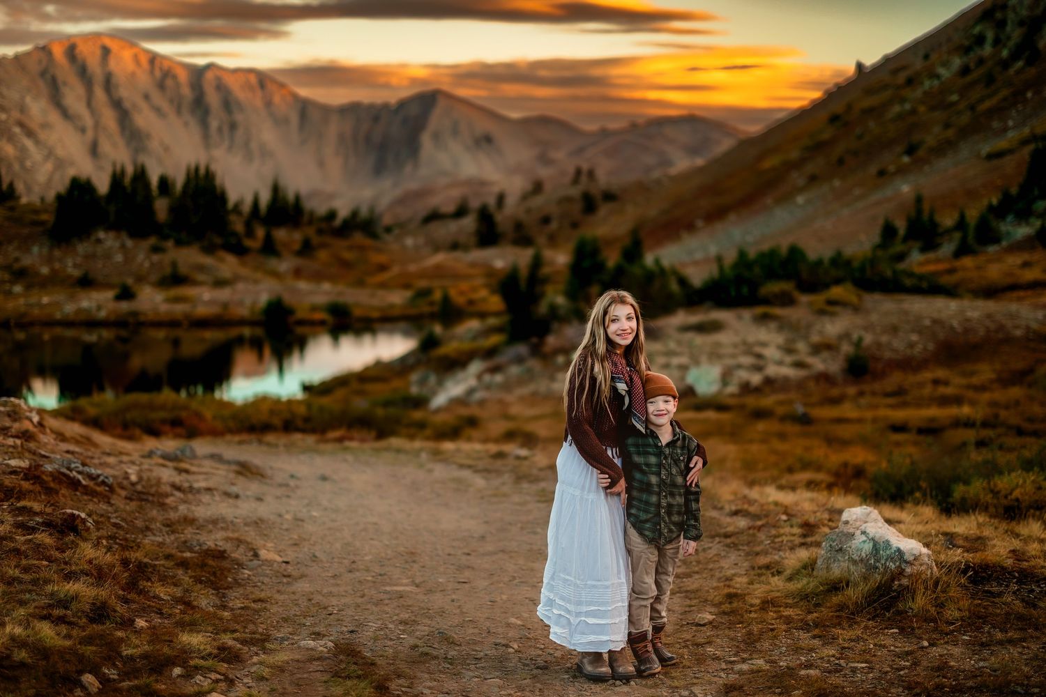 A woman and young boy stand on a mountain trail at sunset with a reflective lake and dramatic sky behind them.