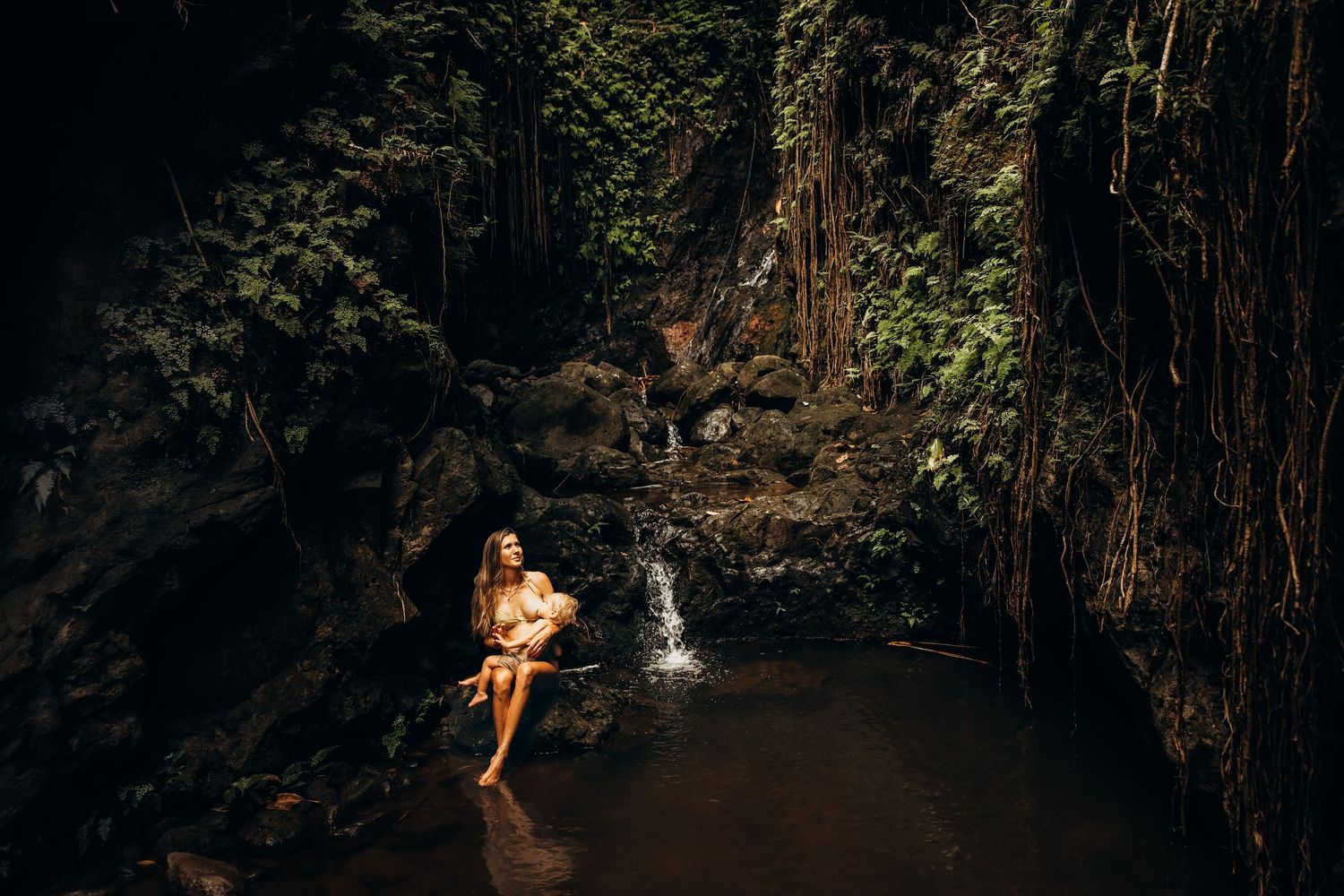 Woman sitting by a serene jungle waterfall surrounded by lush green tropical foliage and dark rocky walls.