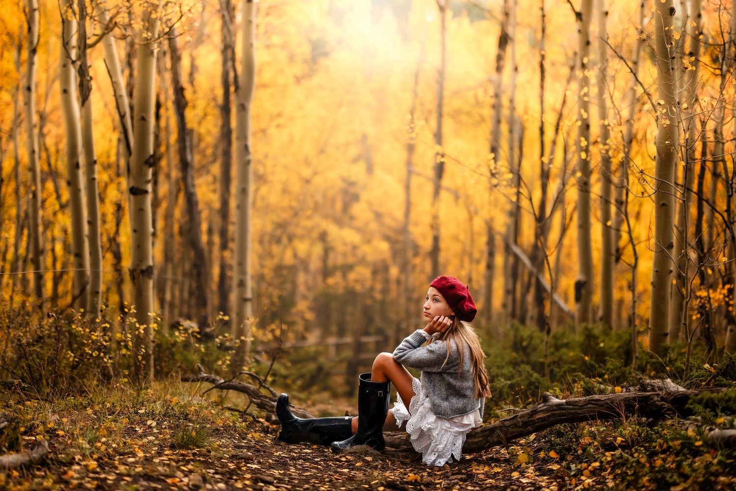 Woman in red beret sits on fallen log in golden autumn aspen forest, surrounded by yellow fall foliage.