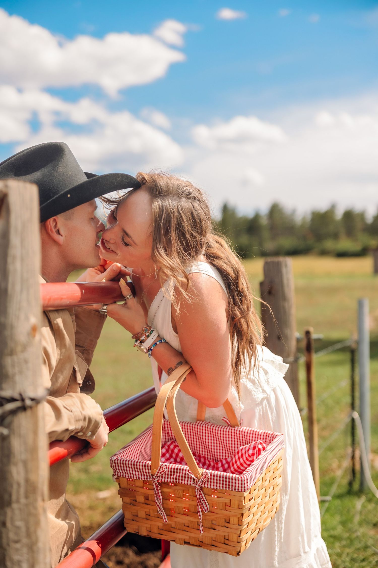 Couple sharing a romantic kiss by a rustic fence, woman in white dress holding a picnic basket on a sunny farm day.