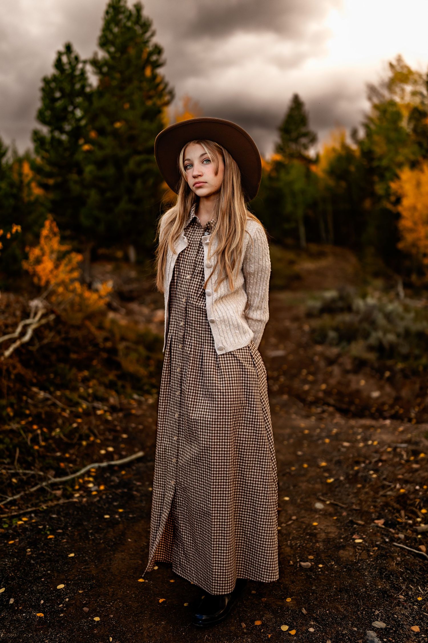 Young woman in plaid maxi coat and brown hat poses on autumn forest path with dramatic stormy sky.