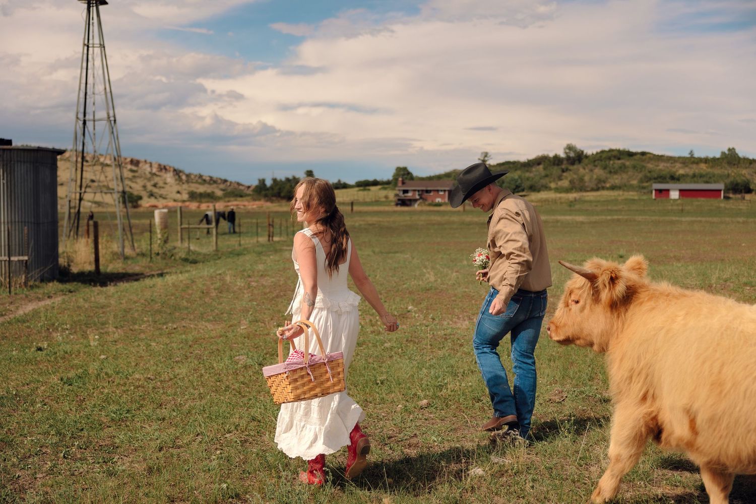 A couple walks toward a fluffy Highland cow on a green farm with a windmill and red barn under cloudy skies.