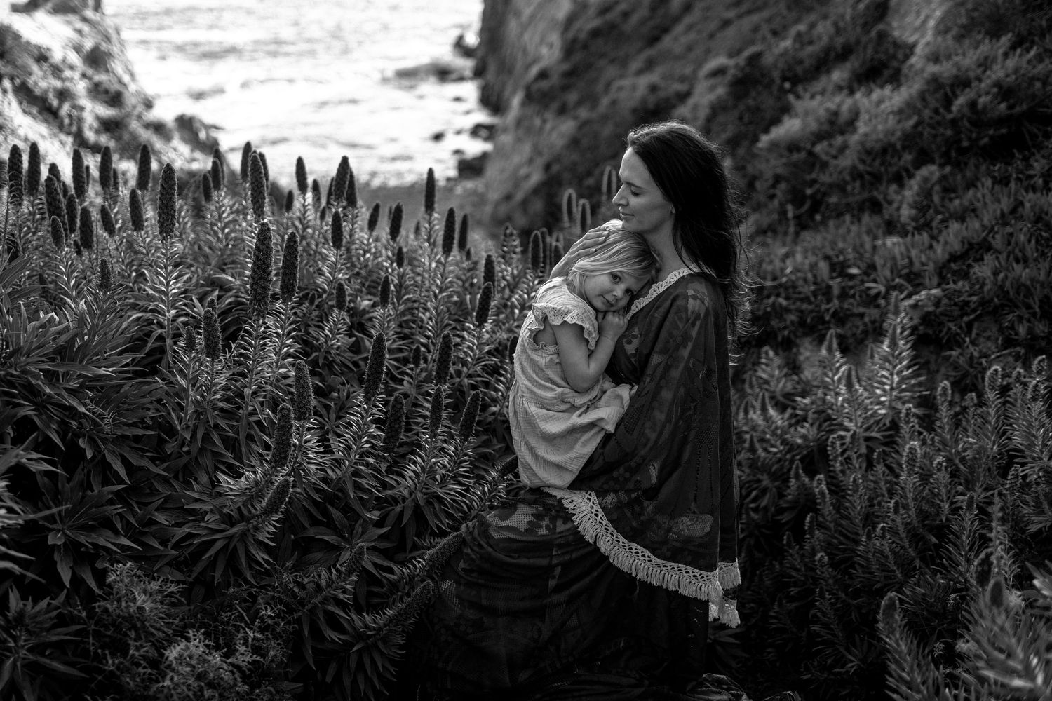 Black and white photo of a mother cradling her baby among tall wildflowers near a rocky coastal cliff.