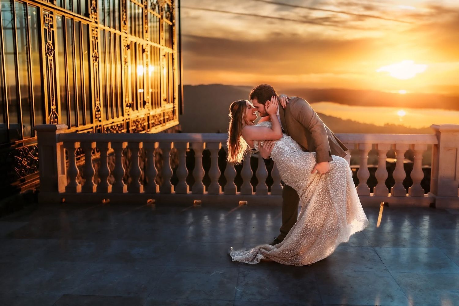 Bride and groom share a romantic dip kiss on a balcony at golden sunset with ocean views.