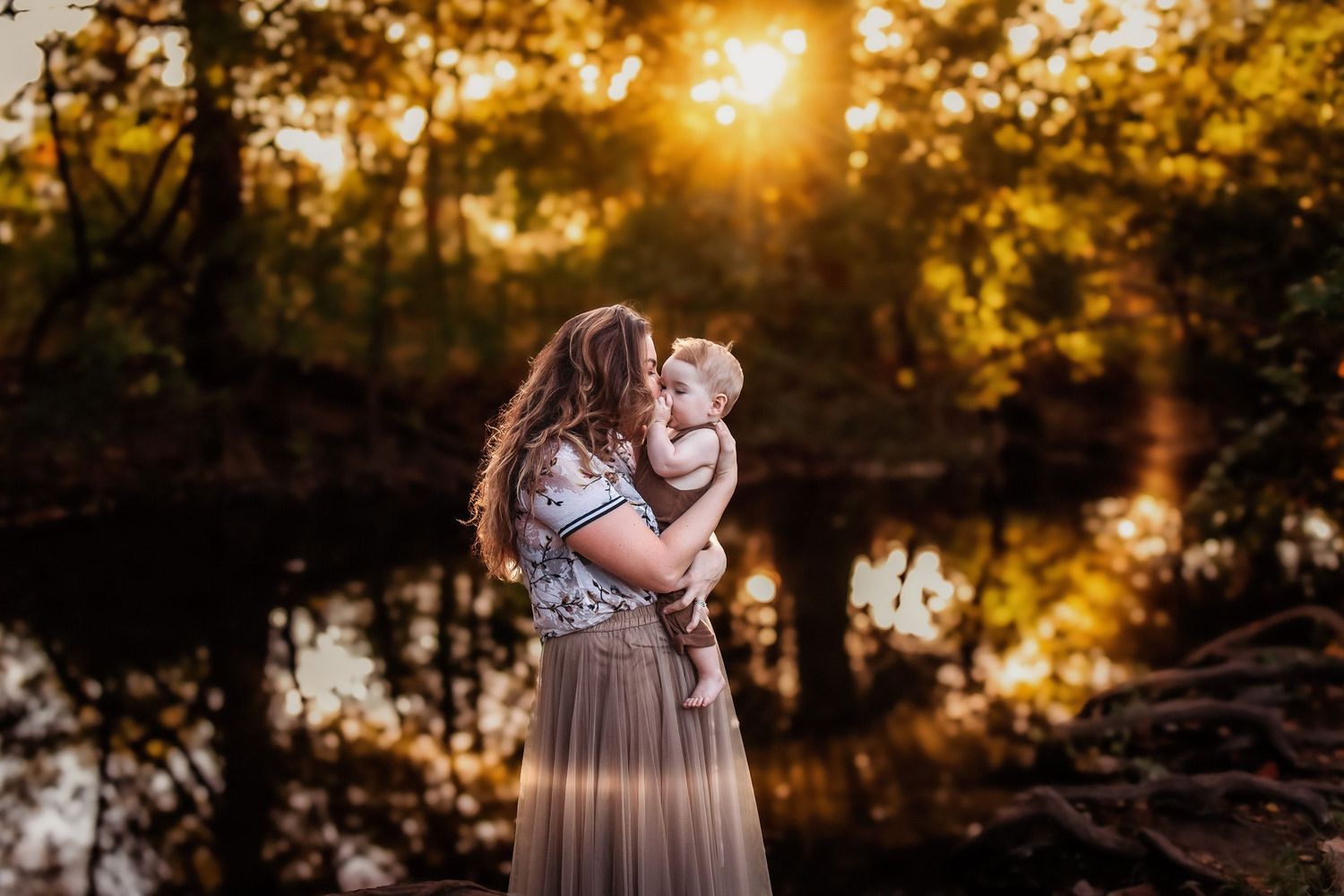 Mother holding baby in golden sunset light near a reflective pond surrounded by lush autumn trees.