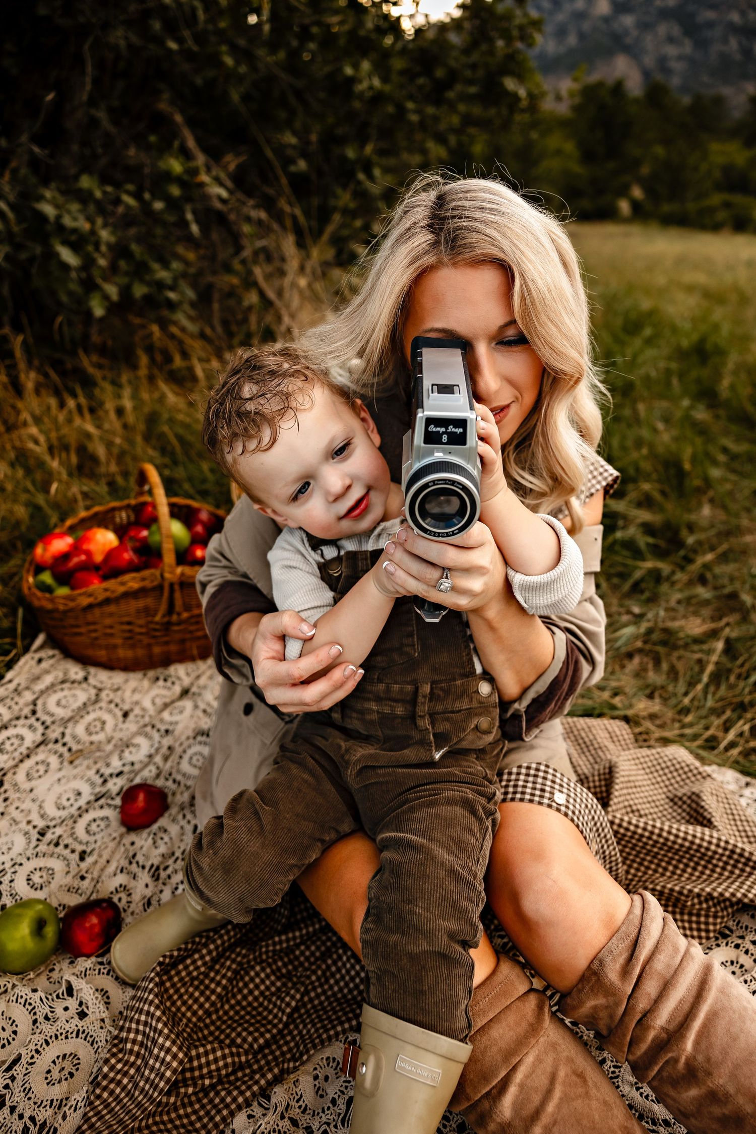 Smiling woman and toddler boy on a picnic blanket holding a vintage film camera, with a basket of red apples and a grassy field behind them.
