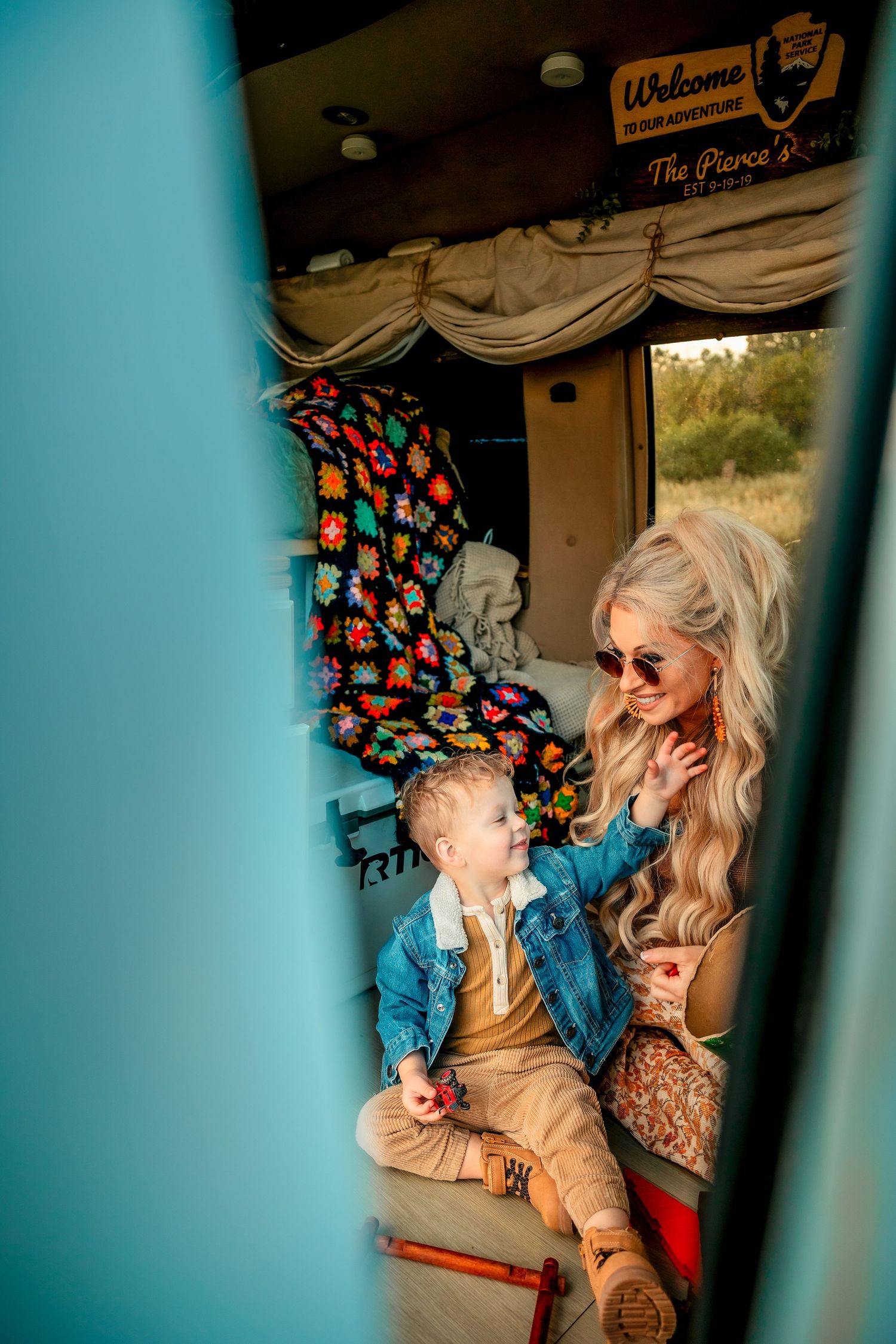 A blonde woman in sunglasses sits with a toddler inside a vintage camper van decorated with a colorful crochet blanket.