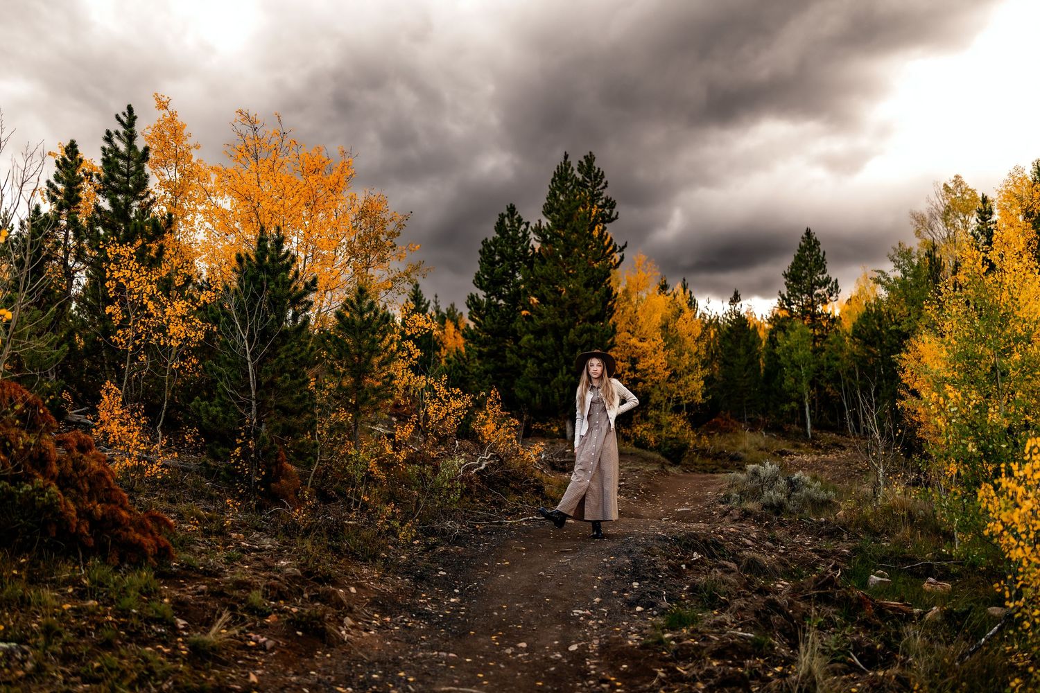 Woman in white dress stands on a forest path surrounded by vibrant autumn foliage under dramatic stormy skies.