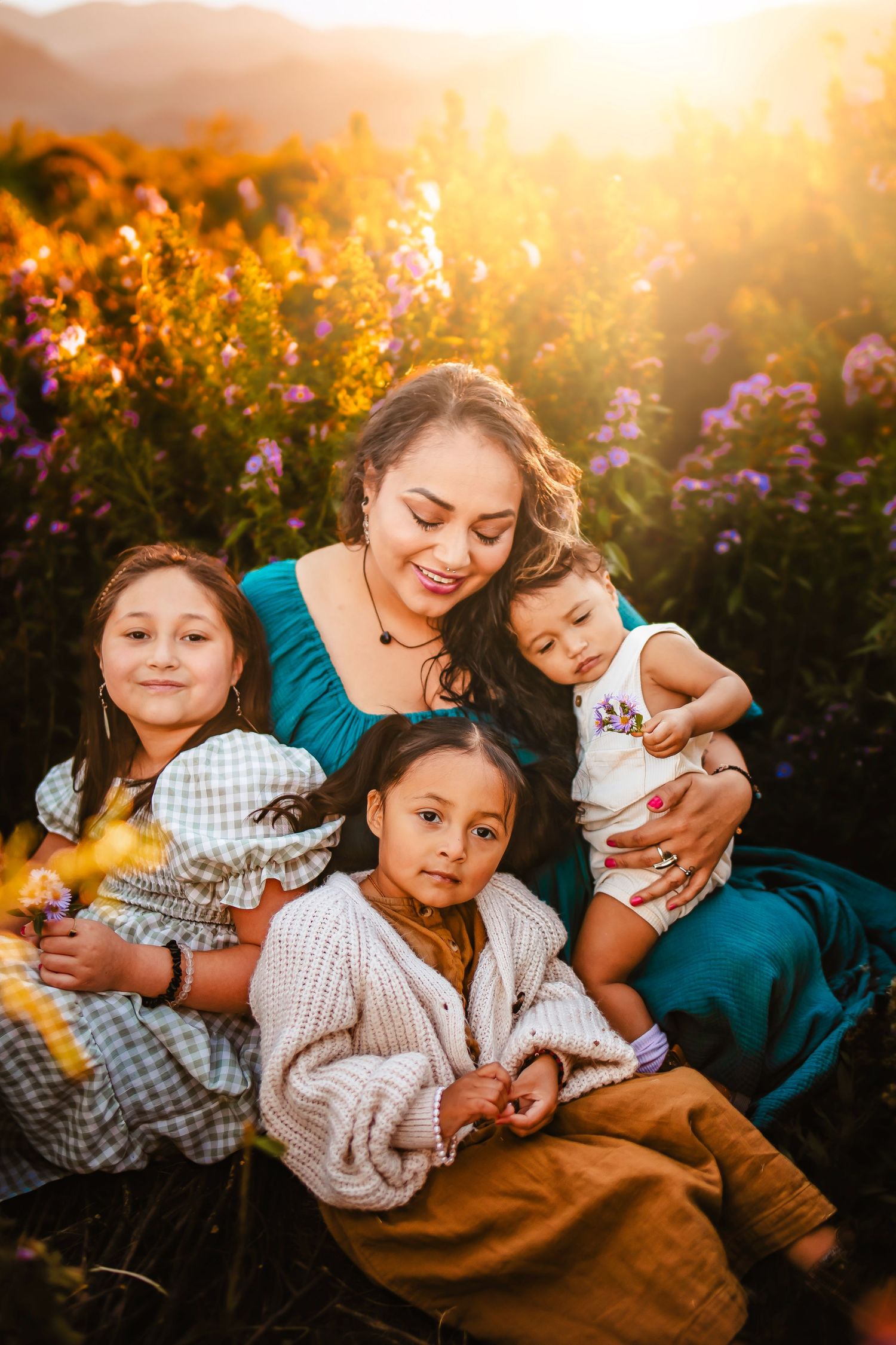 A mother smiles warmly while holding three young children in a sunlit wildflower field at golden hour.