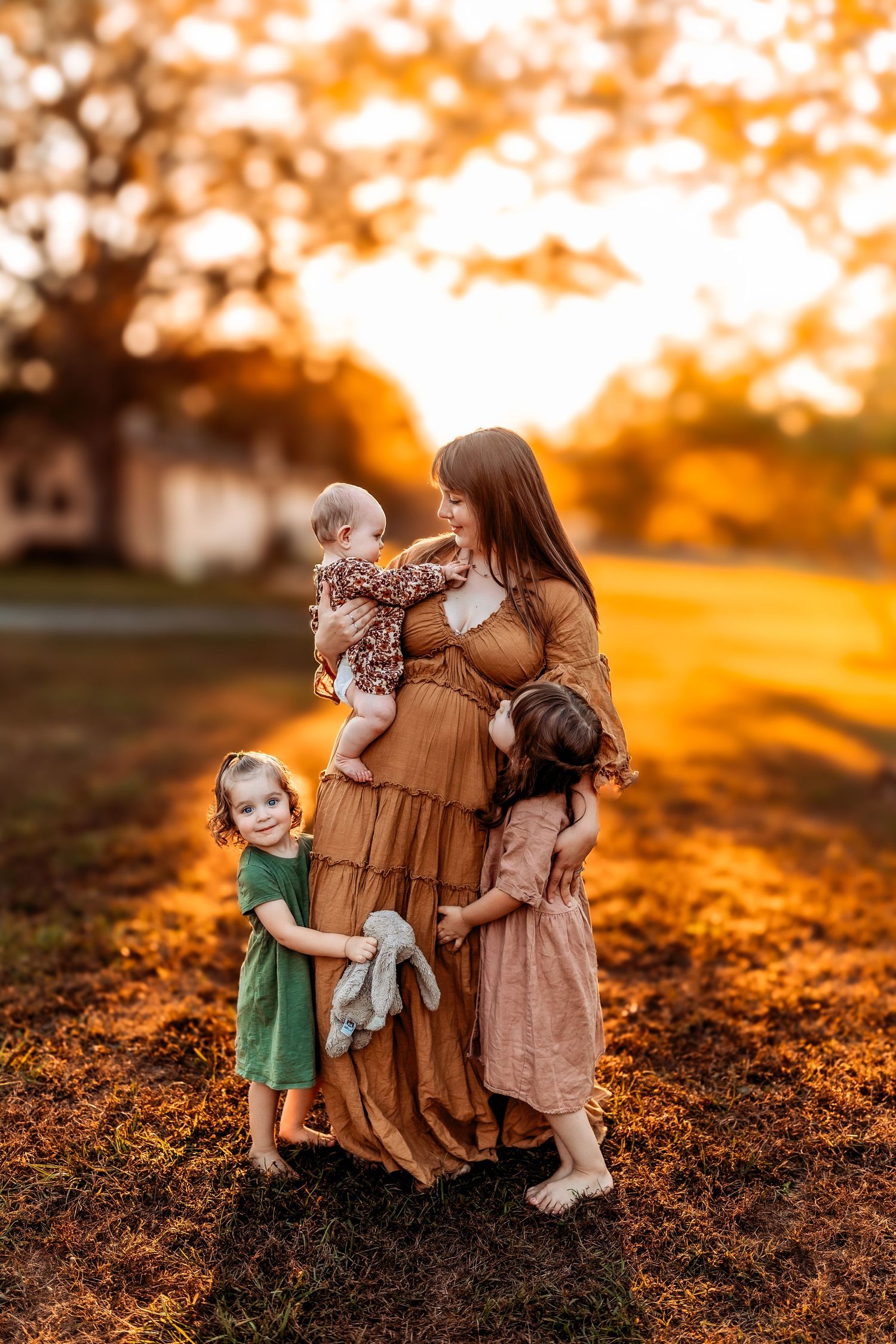 Mother in golden dress holds baby while two young daughters embrace her at sunset in a warm autumn outdoor setting.