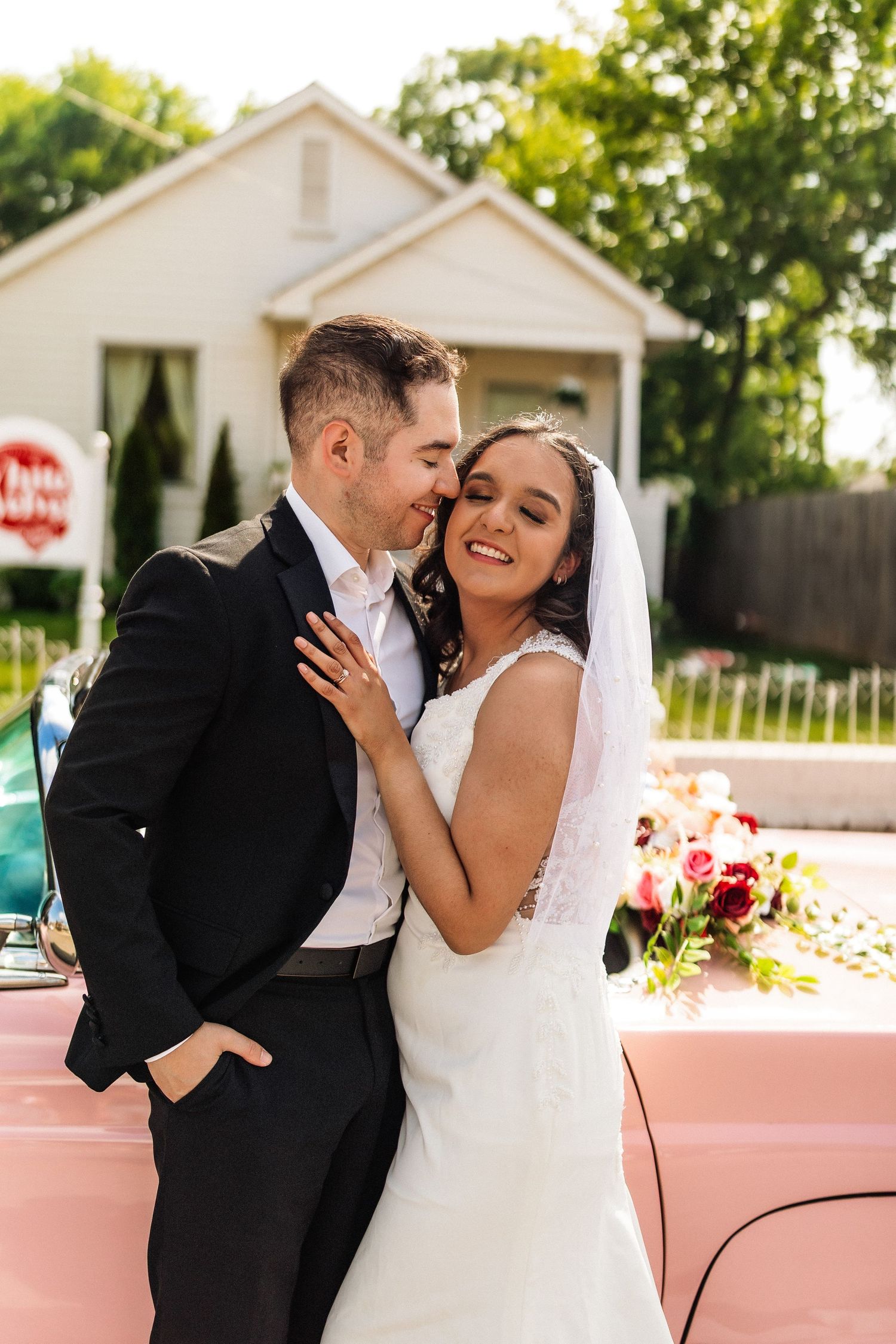 Bride and groom embracing beside a pink vintage car, smiling joyfully in wedding attire outdoors.