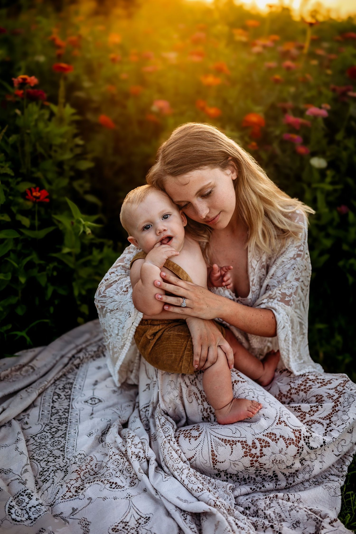 Mother tenderly holds smiling baby in flower field at golden hour sunset, wrapped in boho floral blanket.