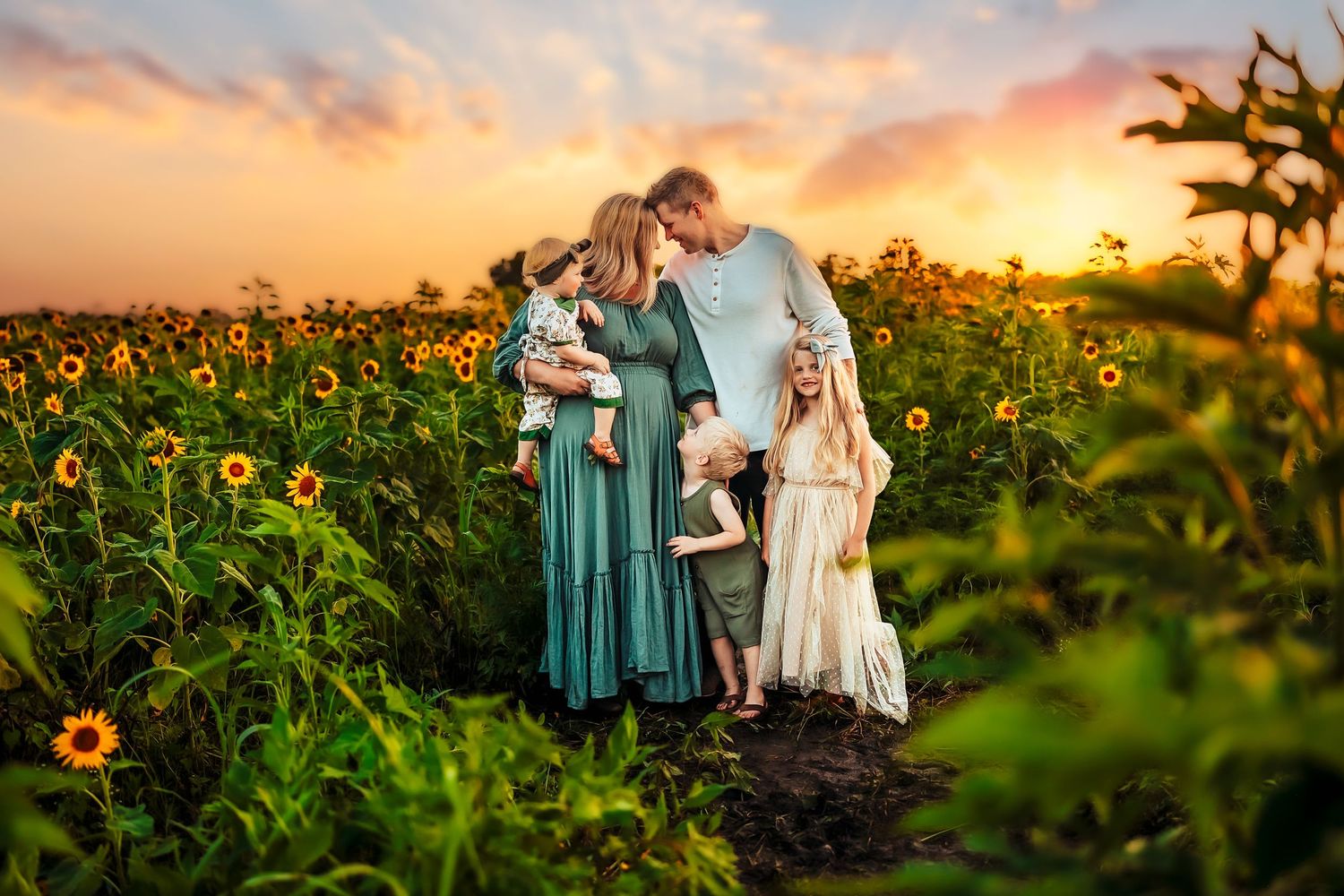 Family posing in sunflower field at golden sunset, parents holding children in teal and white dresses.