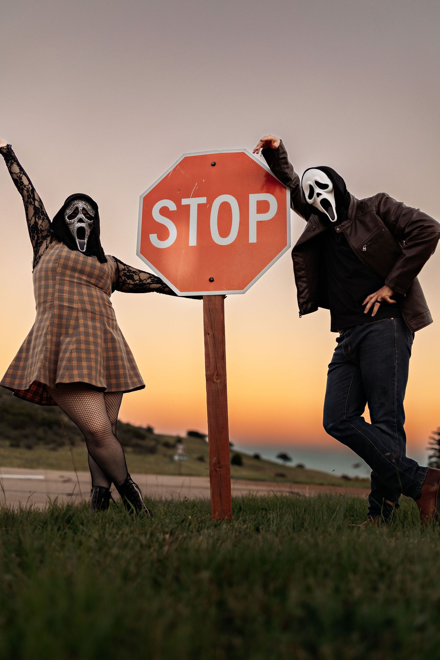 Two people in Ghostface masks pose beside a red stop sign on a grassy hill at sunset, one in a plaid dress and one in a leather jacket.
