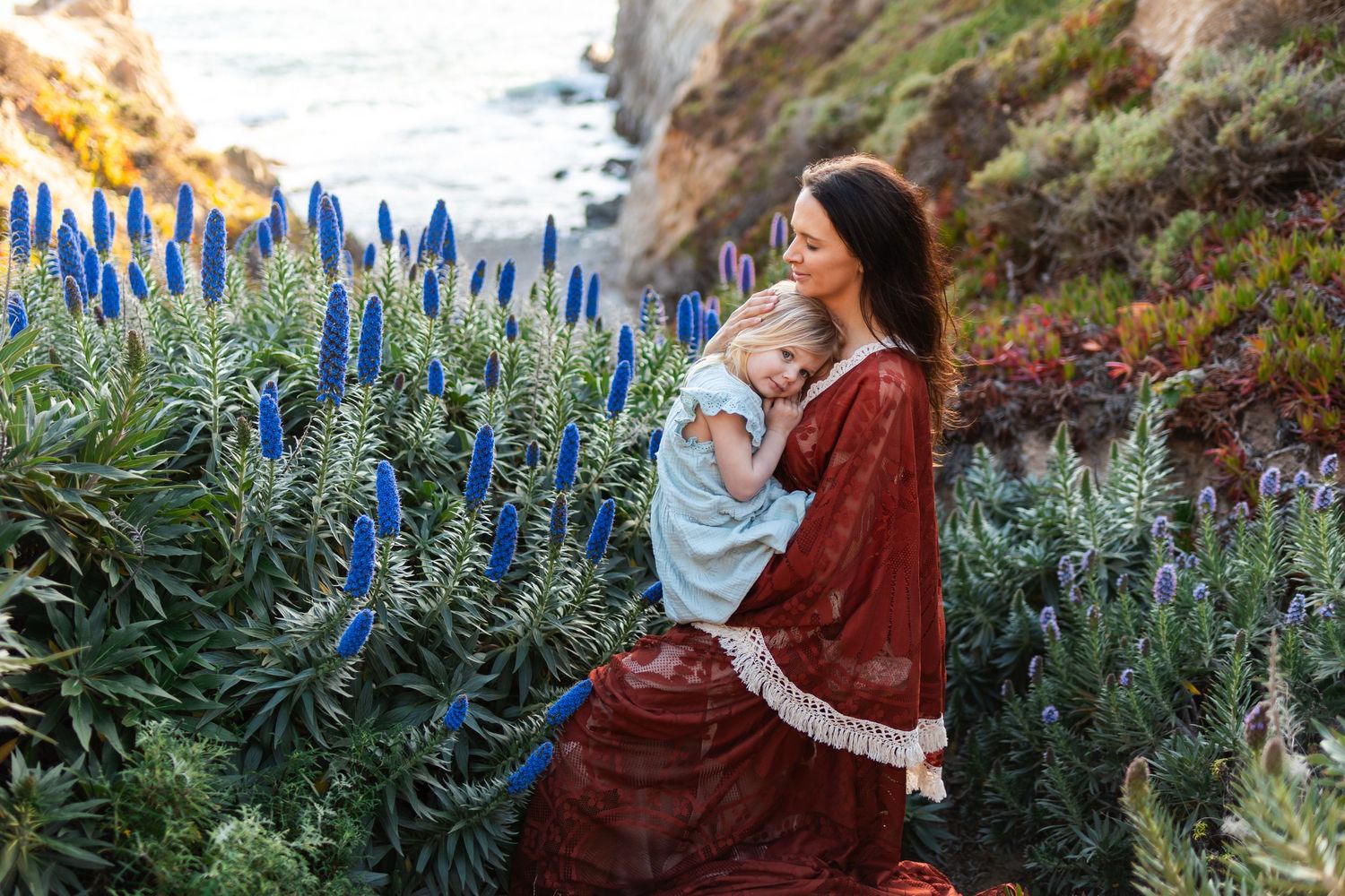 Woman in red dress holds a child among blue Pride of Madeira flowers near a coastal cliff.