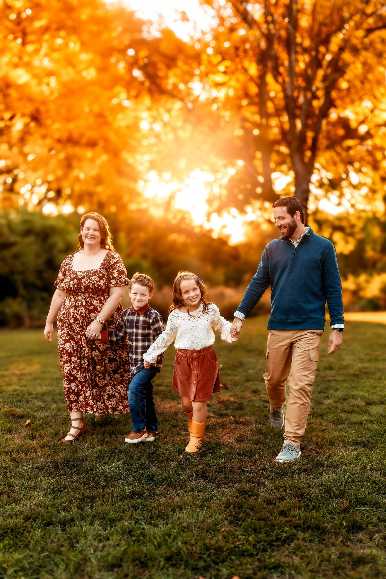 Family of four walking hand-in-hand through a park with vibrant autumn foliage glowing golden in the sunset.