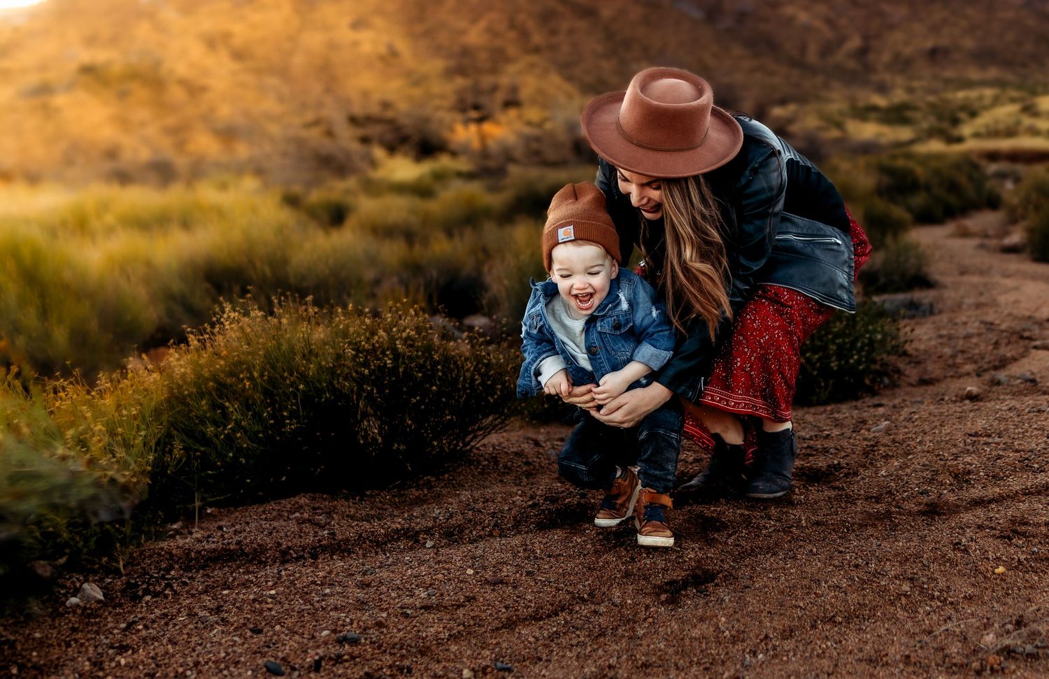 A mother and toddler crouch together on a dirt path, smiling, surrounded by golden desert shrubs at sunset.
