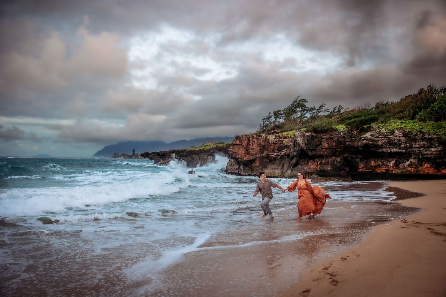 Couple holding hands running along a dramatic Hawaiian beach with stormy skies and crashing waves.