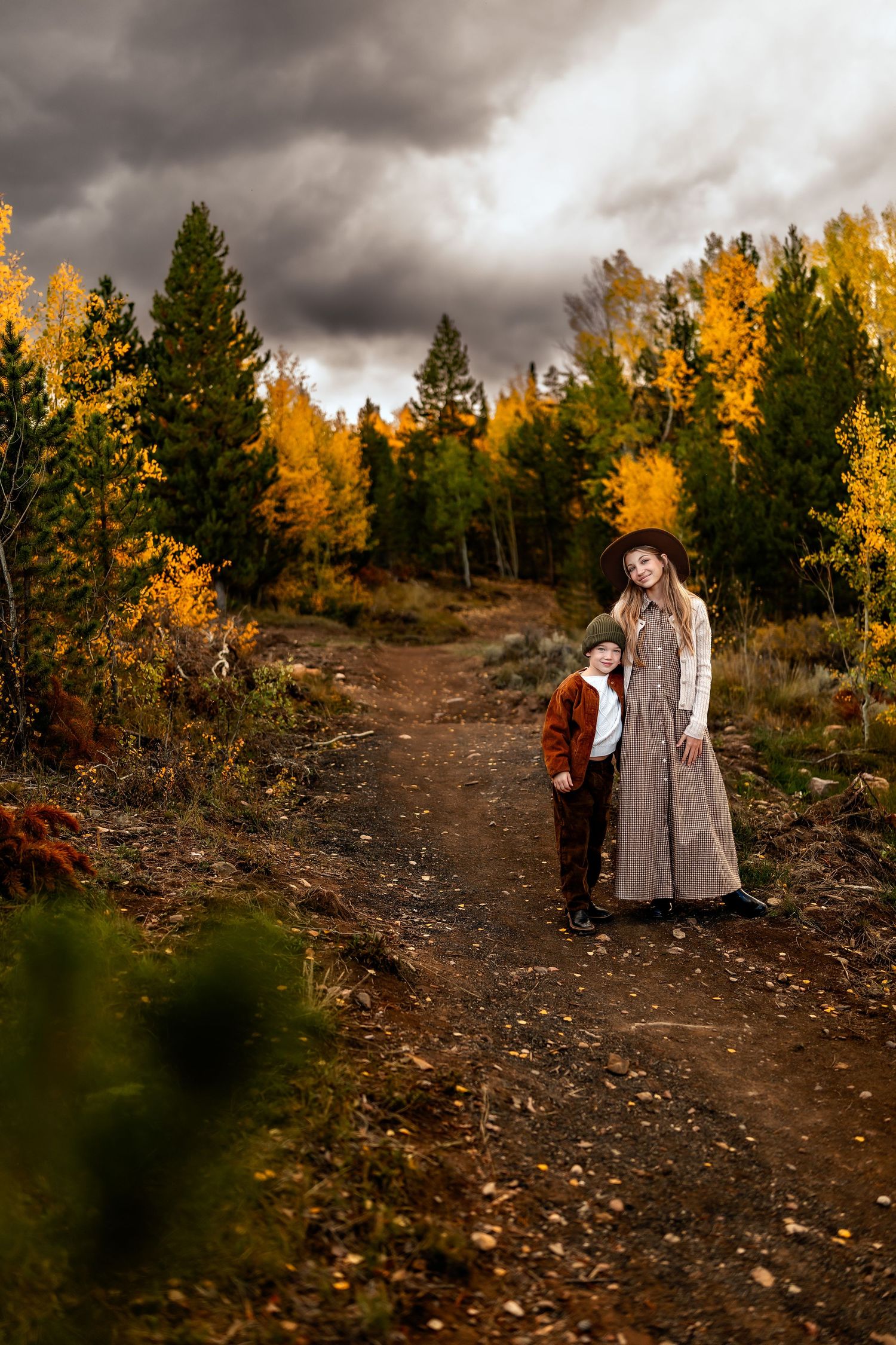 A woman in a striped dress and hat stands with a young boy on an autumn forest path under dramatic stormy skies.