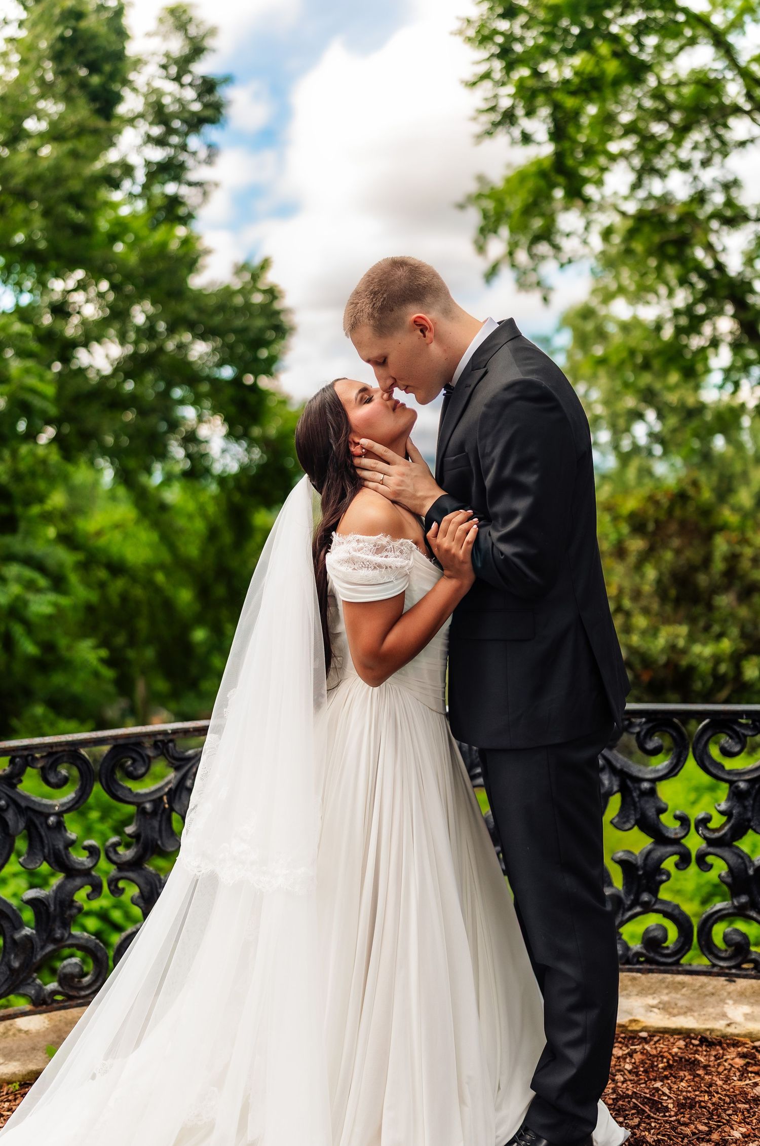 Bride and groom sharing a romantic kiss in a lush garden, she wearing a white off-shoulder gown with veil.