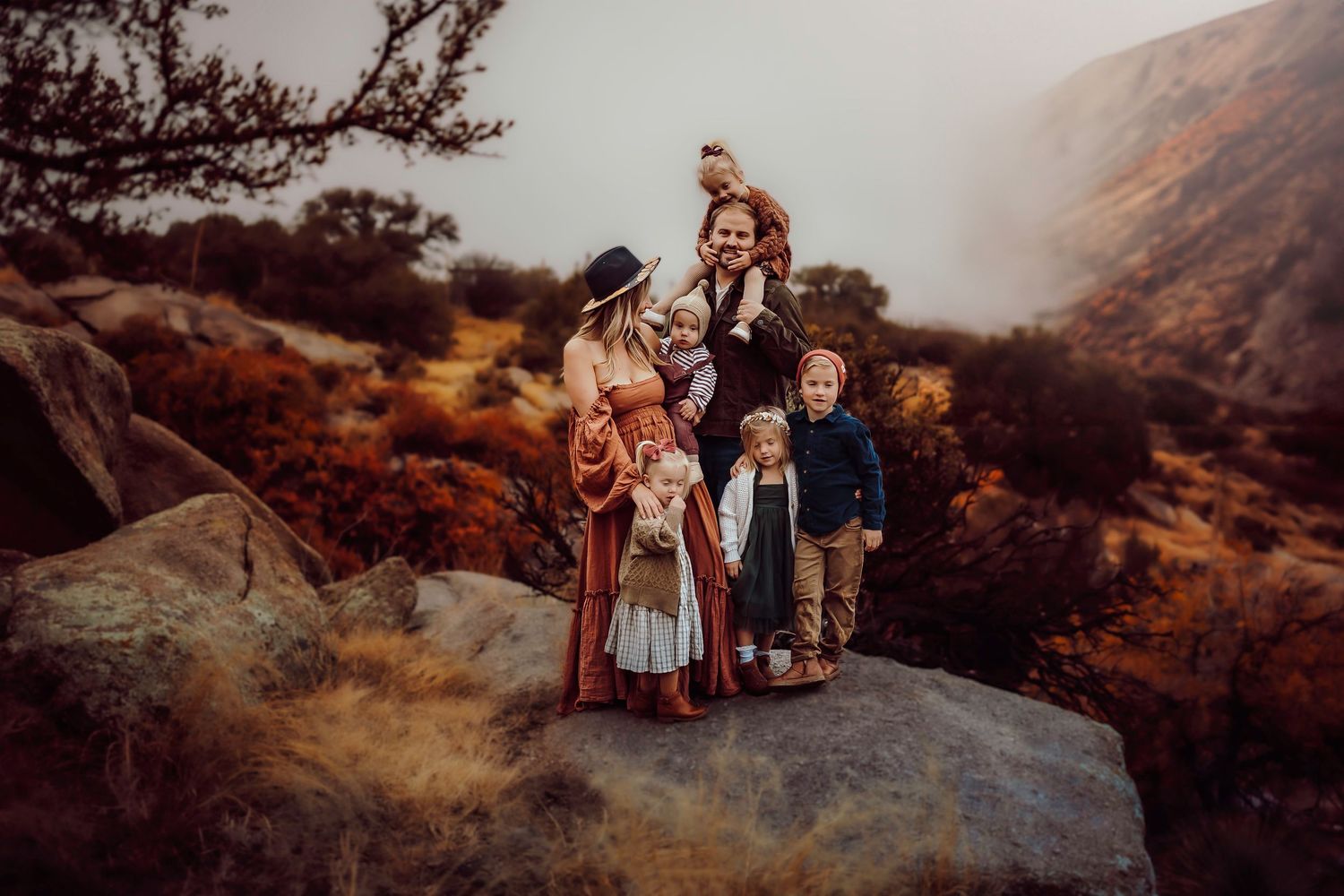 Family posing on rocky outcrop in autumn mountain landscape, wearing rustic bohemian clothing in warm earthy tones.