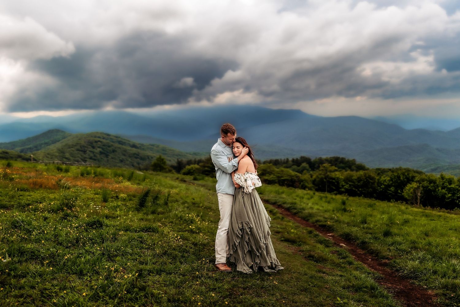 Couple embraces on a mountain meadow in dramatic stormy skies, bride in gray gown holding bouquet.