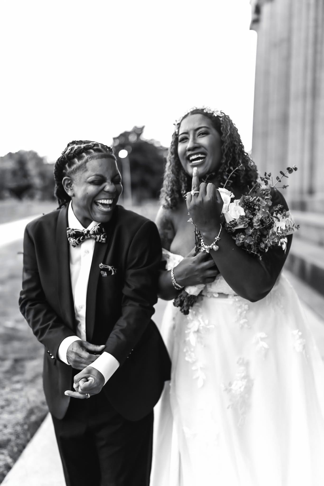 Two brides laugh joyfully outdoors in black and white wedding portrait.