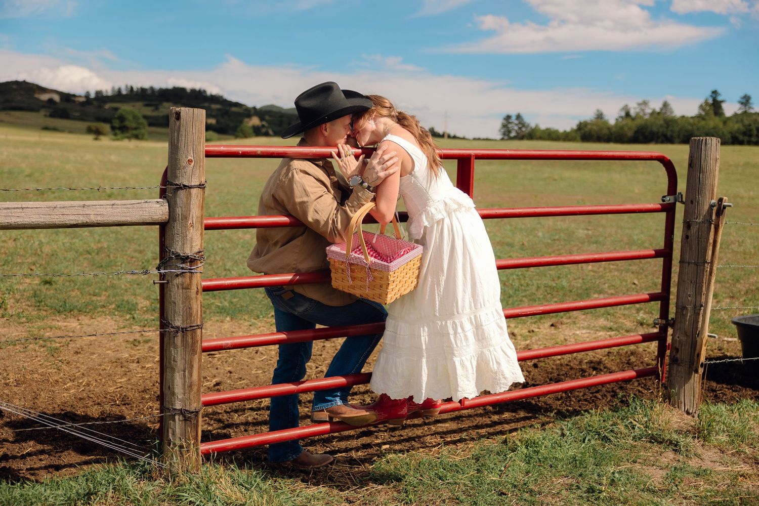 A couple shares a romantic kiss over a red farm gate in a lush green field, with the woman in a white dress.