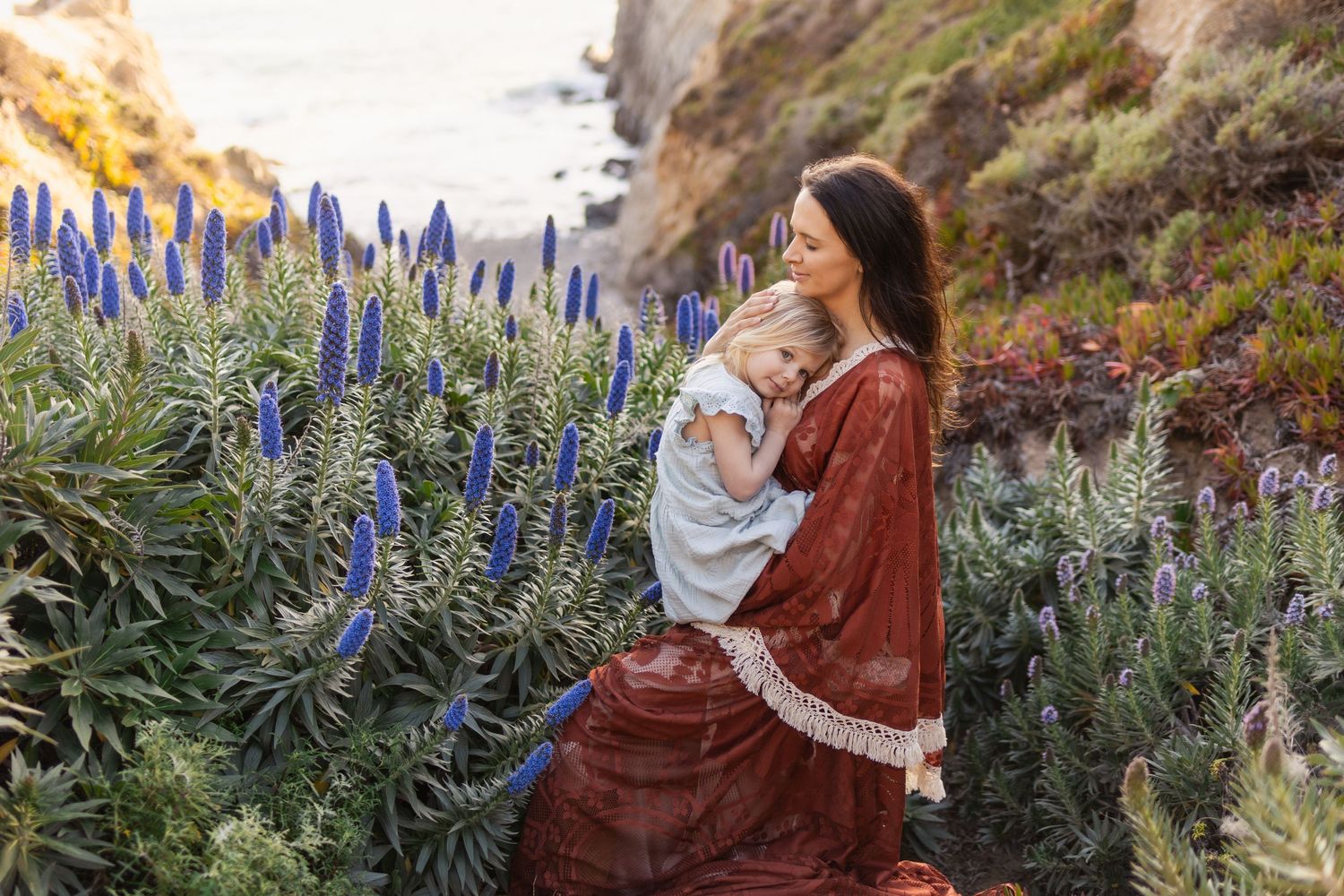 Mother in red dress holds baby among blue wildflowers on coastal cliffside at golden hour.