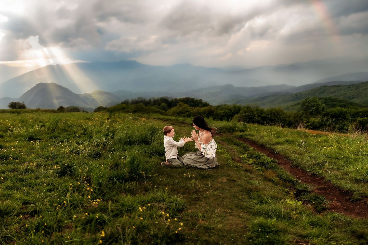 Couple kneeling in green meadow with rainbow and dramatic sunrays over misty mountains in background.