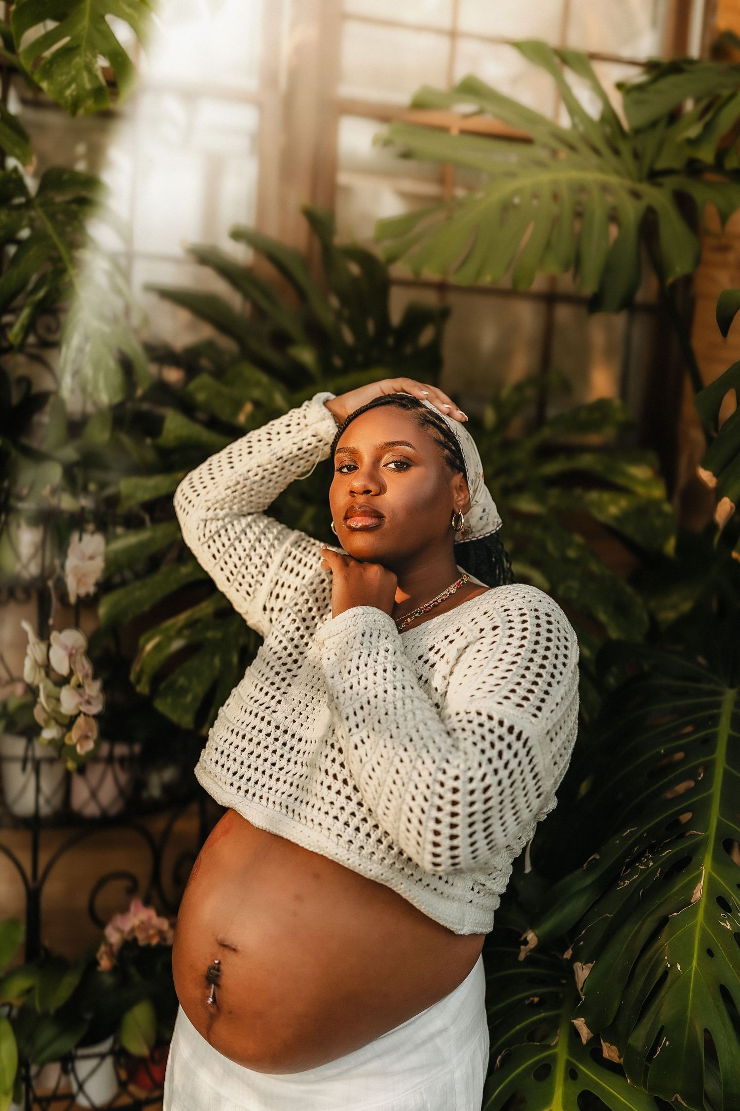 Pregnant Black woman in white crochet crop top poses gracefully among lush tropical plants in warm golden light.