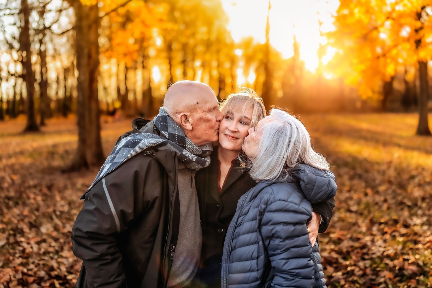 Two elderly people kissing the cheeks of a smiling woman in a sunny autumn park with golden foliage.