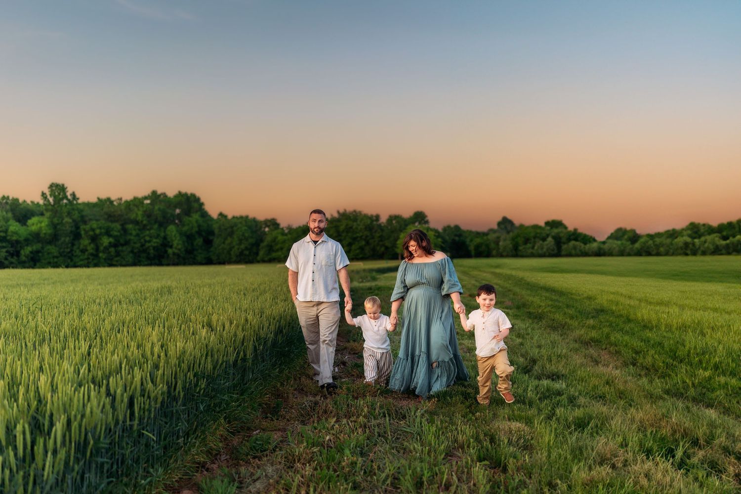 Family of four walking hand-in-hand through a lush green field at golden hour sunset.