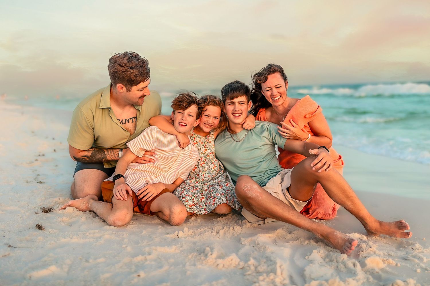 Happy family of five sitting together on a sandy beach at sunset, smiling and embracing near ocean waves.